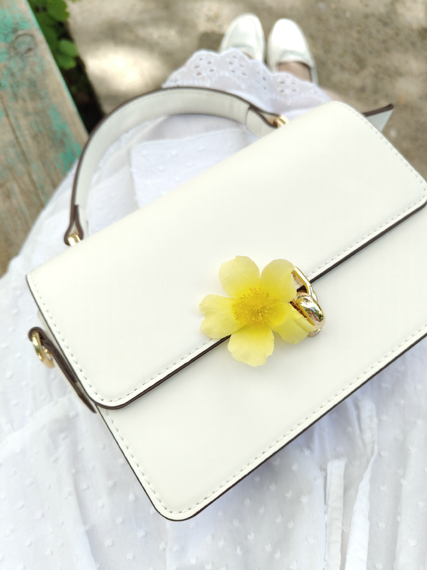 The image presents a close-up, high-angle view of a white handbag, adorned with a yellow flower near its golden clasp. The bag is positioned on a white fabric, possibly a dress. Above the handbag are a pair of white shoes, suggesting the wearer is seated. In the upper left corner, a portion of a green and weathered bench can be observed. The location appears to be an outdoor setting, likely a park or garden.