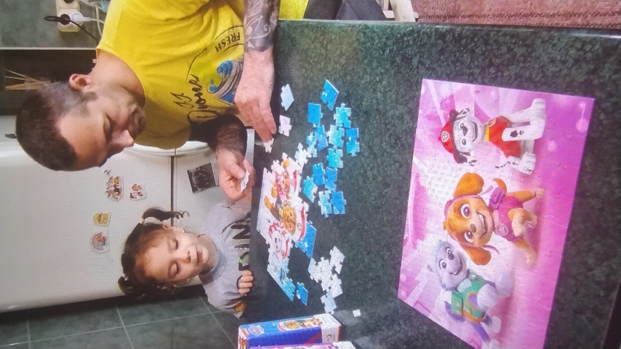 This media file shows a father and daughter working on a jigsaw puzzle at a table. The father, wearing a yellow t-shirt with text partially visible, is focused on the puzzle. The daughter, lying on her side, appears to be concentrating on the puzzle pieces as well. The puzzle depicts characters from Paw Patrol, including Marshall, Skye, and Rocky, on a pink background. Scattered puzzle pieces are spread across the dark-colored tabletop, with a majority of blue and white pieces.
In the background, a refrigerator with several colorful magnets is visible, along with a glimpse of a kitchen area. The lighting suggests it might be daytime. The overall scene conveys a sense of family activity and shared engagement. The location is Mykolaiv, Ukraine.