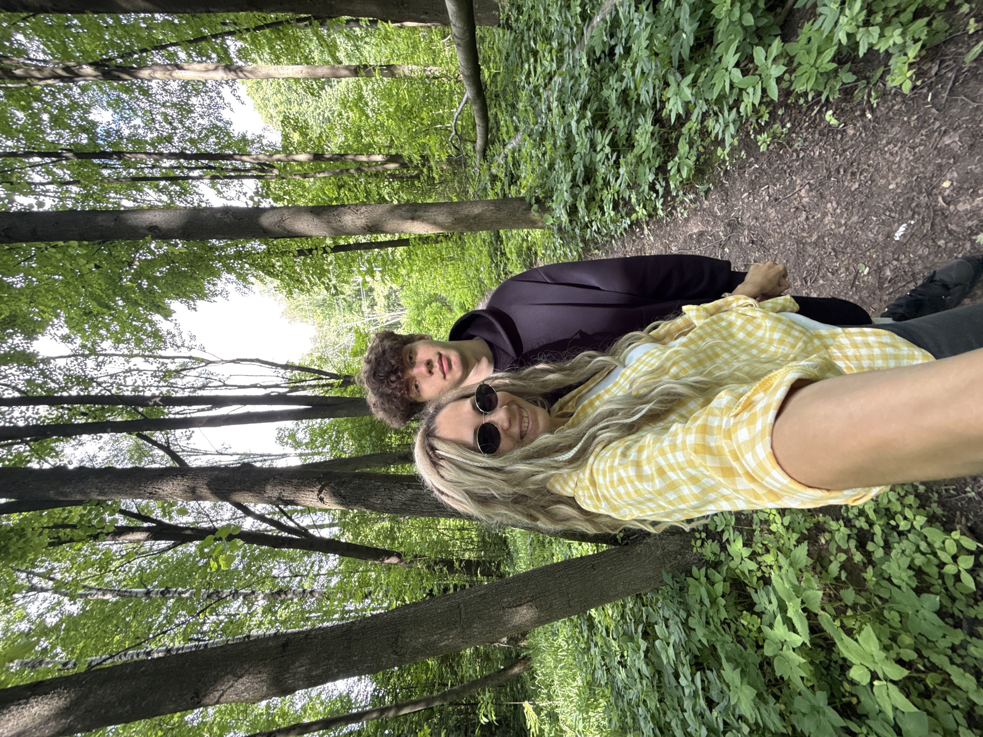 A vertical outdoor shot captures two young individuals in a lush green forest, likely in Rusanovo, Russia. The scene is dominated by tall, slender tree trunks with vibrant green foliage overhead, suggesting a daytime setting with sunlight filtering through the canopy.
In the foreground, a young woman with long, wavy blonde hair, styled with several braids, smiles directly at the camera. She wears round dark sunglasses and a yellow and white plaid shirt. Her arm, possibly holding the camera for a selfie, is visible in the bottom right. Beside and slightly behind her is a young man with curly brown hair, dressed in a dark purple hoodie, looking towards the camera with a neutral expression.
The setting features a clear dirt path to the right, bordered by dense green undergrowth. The forest floor is covered with various plants and leaves. The background shows more trees and bright patches of sky visible through the leaves, indicating an open canopy. The overall atmosphere suggests a pleasant day for an outing or hike in nature. No visible text is present in the image.