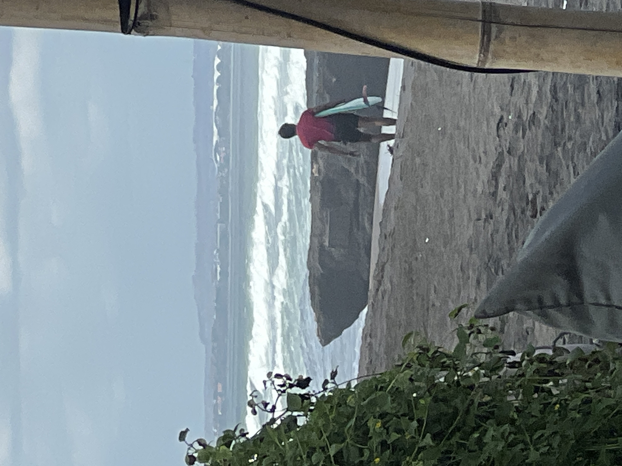 A surfer in a red t-shirt and black shorts walks towards the ocean on a dark sand beach in North Kuta, Indonesia. The surfer carries a light blue surfboard under their right arm. In the foreground, lush green foliage obstructs the view on the left, and a dark grey beanbag chair partially fills the bottom of the frame. The ocean is choppy with white waves crashing against dark rock formations and a distant shoreline with buildings. The sky is overcast and pale blue. A thick bamboo pole is visible on the right side of the image.