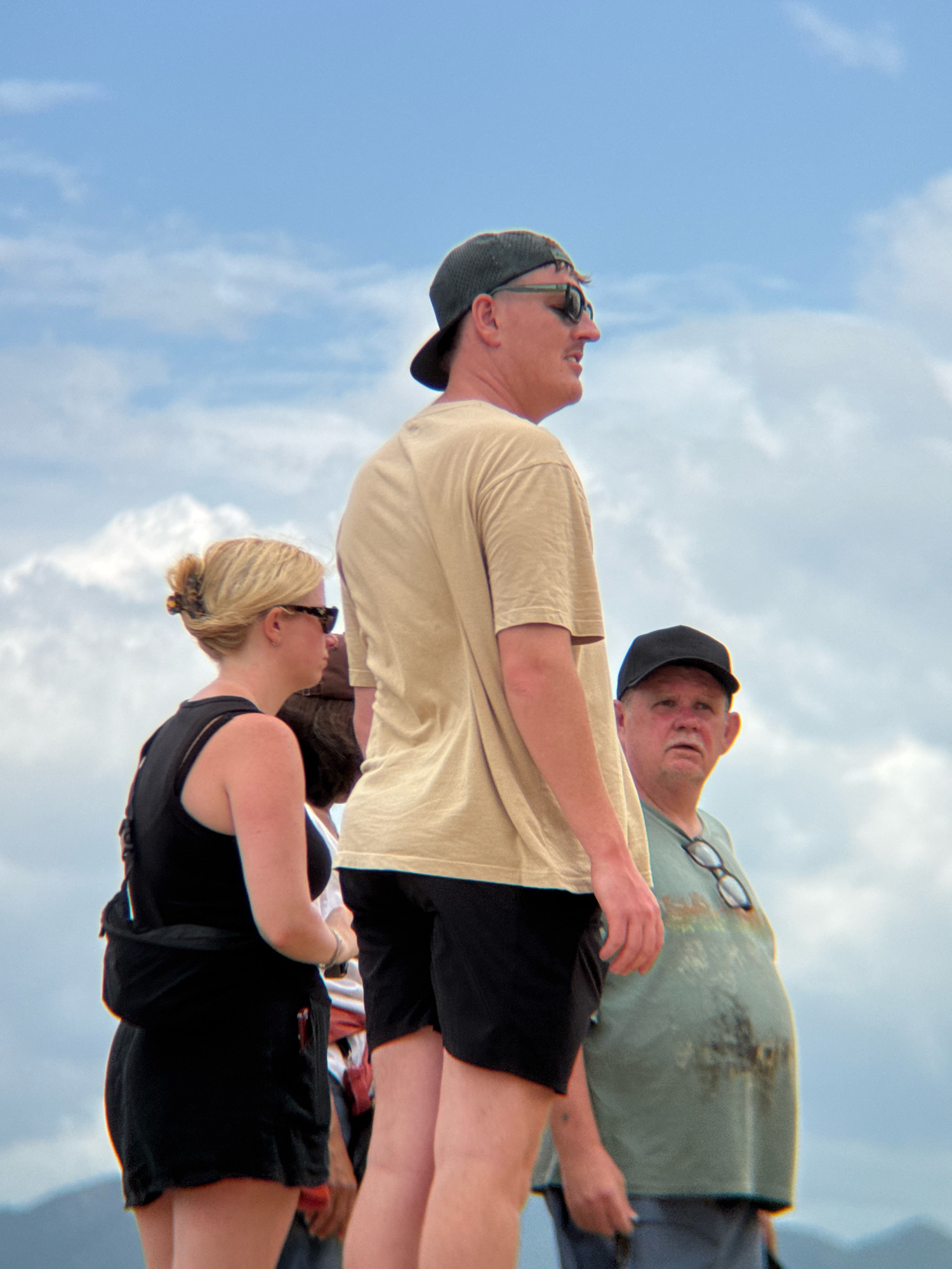 A group of three adults, two males and one female, are pictured outdoors against a cloudy sky, with mountains visible in the distance. The central subject is a male wearing a tan t-shirt, black shorts, a dark baseball cap, and sunglasses, looking to his right. To his left, a female with blonde hair pulled back in a bun, wearing a black tank top and black shorts, looks to her left. Behind her, a partial view of another person is visible. To the right of the central subject, an older male wearing a light green t-shirt with a graphic print, dark pants, and a black baseball cap, is looking towards the viewer with a concerned expression. This scene is likely located in the city of Nha Trang, Vietnam, a coastal resort city known for its beaches and mountainous backdrop.