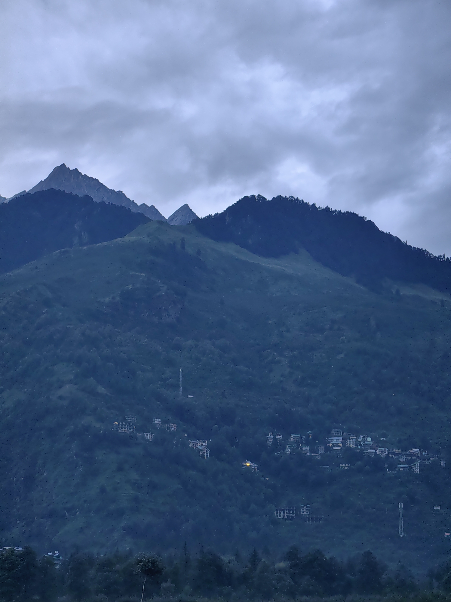 The image depicts a twilight scene of a mountainous landscape with a village nestled on the slopes. The sky is overcast with heavy, dark clouds, creating a somber atmosphere. Jagged mountain peaks, shrouded in shadow, dominate the upper portion of the frame, their rugged contours softened by the diffused light. Below the peaks, the mountainside is covered in dense, dark green vegetation, with patches of lighter green indicating open grassy areas or cultivation.
Scattered across the middle and lower slopes of the mountains are clusters of buildings that form a village. These structures appear to be multi-story, with lighter-colored facades, some of which show illuminated windows, suggesting that night is falling and interior lights are on. The village is interspersed with trees, and a tall, slender telecommunications tower is visible on one of the slopes. The foreground consists of a dark, tree-covered area, possibly a forest or dense shrubbery, which gradually fades into the darker tones of the lower landscape. The overall impression is one of a remote, tranquil settlement at the foot of imposing mountains, under a brooding sky. The location is identified as Bahang, India, a village in the Kullu Valley, Himachal Pradesh, known for its scenic beauty and mountainous terrain.