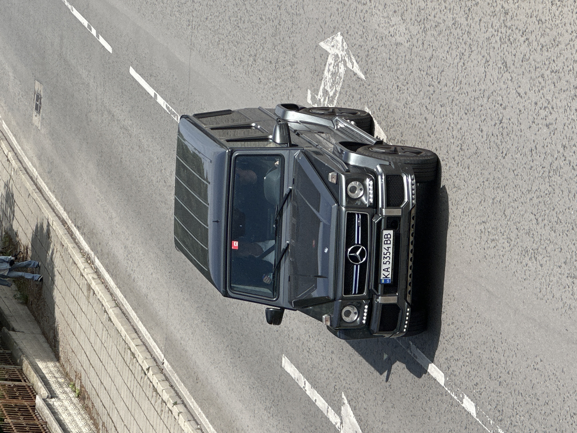 An elevated aerial view captures a gray Mercedes-Benz G-Class SUV on an asphalt road in Veliko Tarnovo, Bulgaria. The car's license plate reads "KA 5354 BB." Road markings include solid and broken white lines defining lanes and a white arrow indicating direction. A concrete curb and wall run along the left side of the road, with a glimpse of a figure in jeans and a jacket standing near a metal grate. The weather appears bright and sunny.