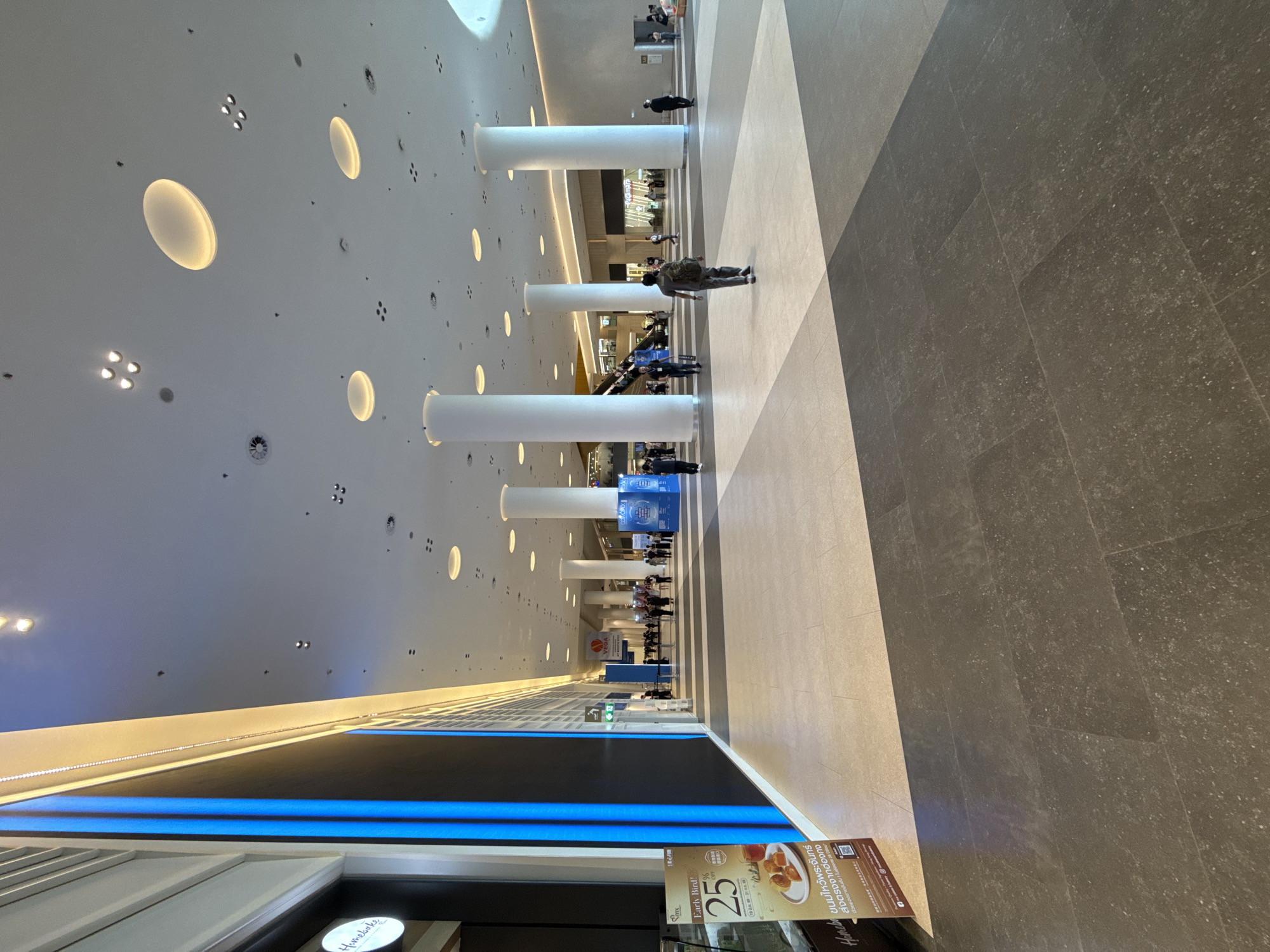 This media file captures a wide-angle view of a modern indoor public space, likely a shopping mall or transit hub, characterized by its expansive white ceiling with embedded oval lights and numerous cylindrical white pillars. The floor is a mosaic of light beige and dark gray tiles arranged in a geometric pattern.
Several people are visible, scattered throughout the space. Some are walking, while others appear to be congregating or waiting. One individual in the foreground is walking away from the camera, carrying a backpack. In the background, more people are seen near escalators and shopfronts, suggesting a busy commercial environment. The lighting is bright and even, indicative of an indoor setting with artificial illumination.
Prominently displayed are various signs and advertisements. One sign, featuring a crescent moon and Chinese characters, announces "ขนมไหว้พระจันทร์" (Mooncakes) and offers an "Early Bird! 25 OFF." Another sign displays "VEGA" and "Pacamara," likely brand names. The overall atmosphere appears neutral, with no strong emotional cues evident. The presence of escalators and the architecture suggest a contemporary urban setting. The visibility of shop facades in the distance hints at the retail nature of the location. The text also indicates it is likely in Thailand, given the Thai language.
