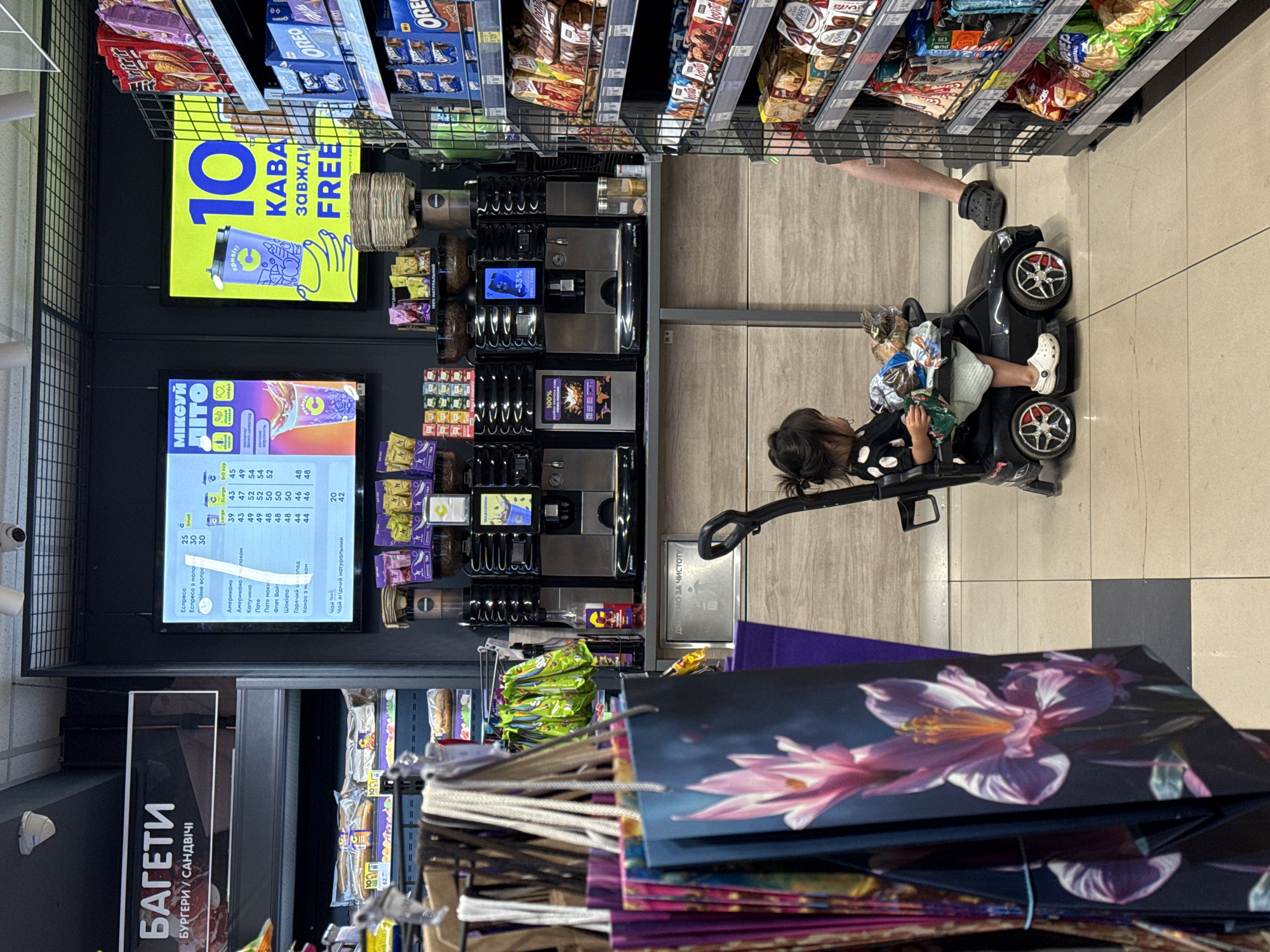 The image captures a scene inside a store, likely a supermarket or convenience store, with a focus on a young child seated in a toy car. The child, dressed in a black and white polka-dot shirt, is holding a green bag of snacks. Next to the child, an adult's legs, wearing black Crocs, are visible, suggesting they are accompanying the child. The store environment is evident with shelves stocked with various food items, including bags of chips and boxes of cookies.
Dominating the background is a large, bright yellow sign with blue text that reads "10 KABA завжди FREE", indicating a promotion for coffee. Below this, a digital screen displays a menu with coffee options and prices, with the text "МІКСУЙ ПІТО" and "100%" visible. A coffee machine is also present, suggesting self-service beverage options.
Further details include stacks of colorful shopping bags in the foreground, some adorned with floral designs, and other shelves displaying merchandise. The lighting appears to be artificial, typical of an indoor retail setting. The overall scene suggests a casual shopping trip, with the child momentarily entertained in the toy car while an adult shops. The presence of Cyrillic text on signs and menus suggests the location is in a country that uses this alphabet, with the provided context indicating Lutsk, Ukraine.