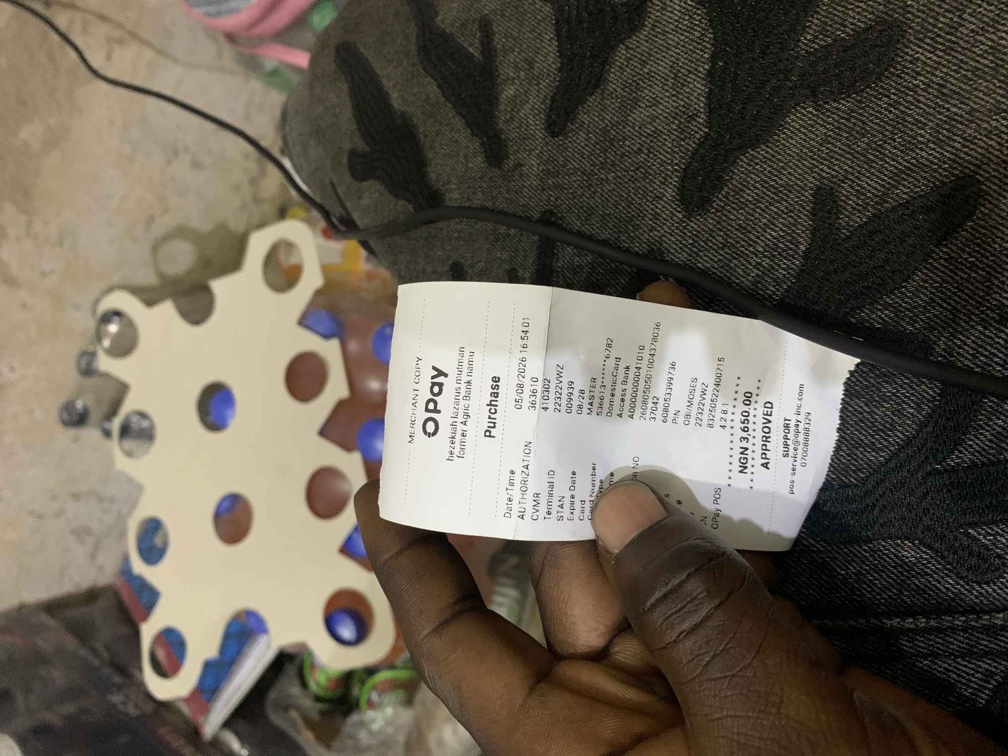 A close-up, waist-up, indoor shot shows a person's right hand, fingers wrapped around a folded paper receipt. The receipt, printed in black text on white paper, displays transaction details. The background is blurred, showing a light-colored, patterned object and dark fabric.
The receipt indicates a purchase made through OPay, with the merchant name "hezekiah lazarus mutman" and "former Agric Bank namu". Transaction details include "Date/Time: 05/08/2026 16:54:01", "AUTHORIZATION: 363610", "CVMR: 540110", "Terminal ID: 410302", "STAN: 22322VWZ", "Expire Date: 08/28", "Card Number: MASTER ******6782", "Card Type: Domestic Card", and "Name: A0000000041010". A PIN is also visible. The transaction amount is listed as "NGN 3,650.00" with the status "APPROVED". Support contact information is provided at the bottom. The image was taken in New Stadium, Nigeria.