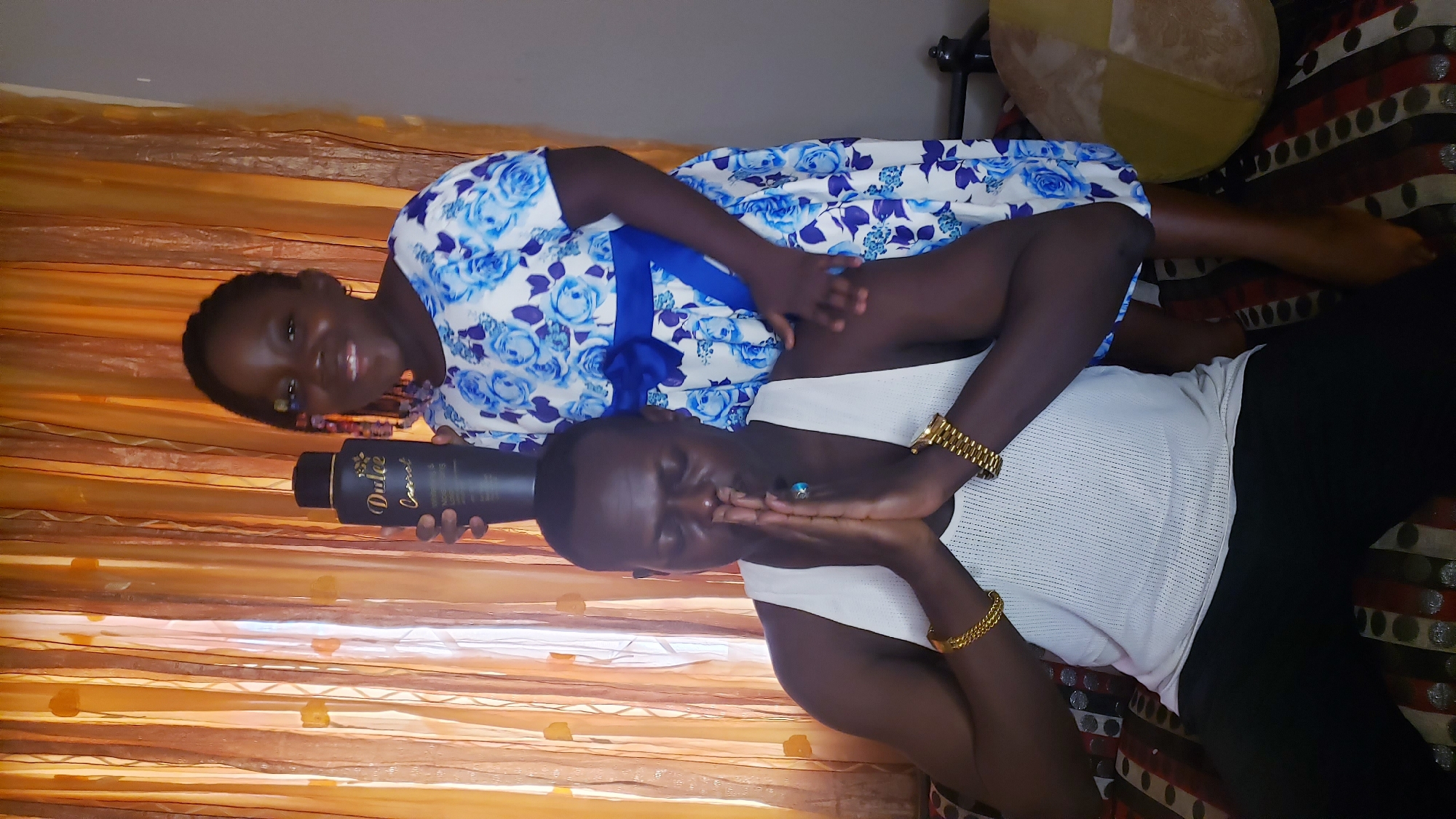 Two people are lying down on a surface. The person on top is a child wearing a blue and white floral dress, holding a black bottle labeled "Dulce." The person below is wearing a white tank top and black pants. Gold bracelets are visible on both wrists. The background includes patterned upholstery and golden-colored curtains. The setting appears to be indoors. No information to confirm the location within Sabon Tasha, Nigeria.