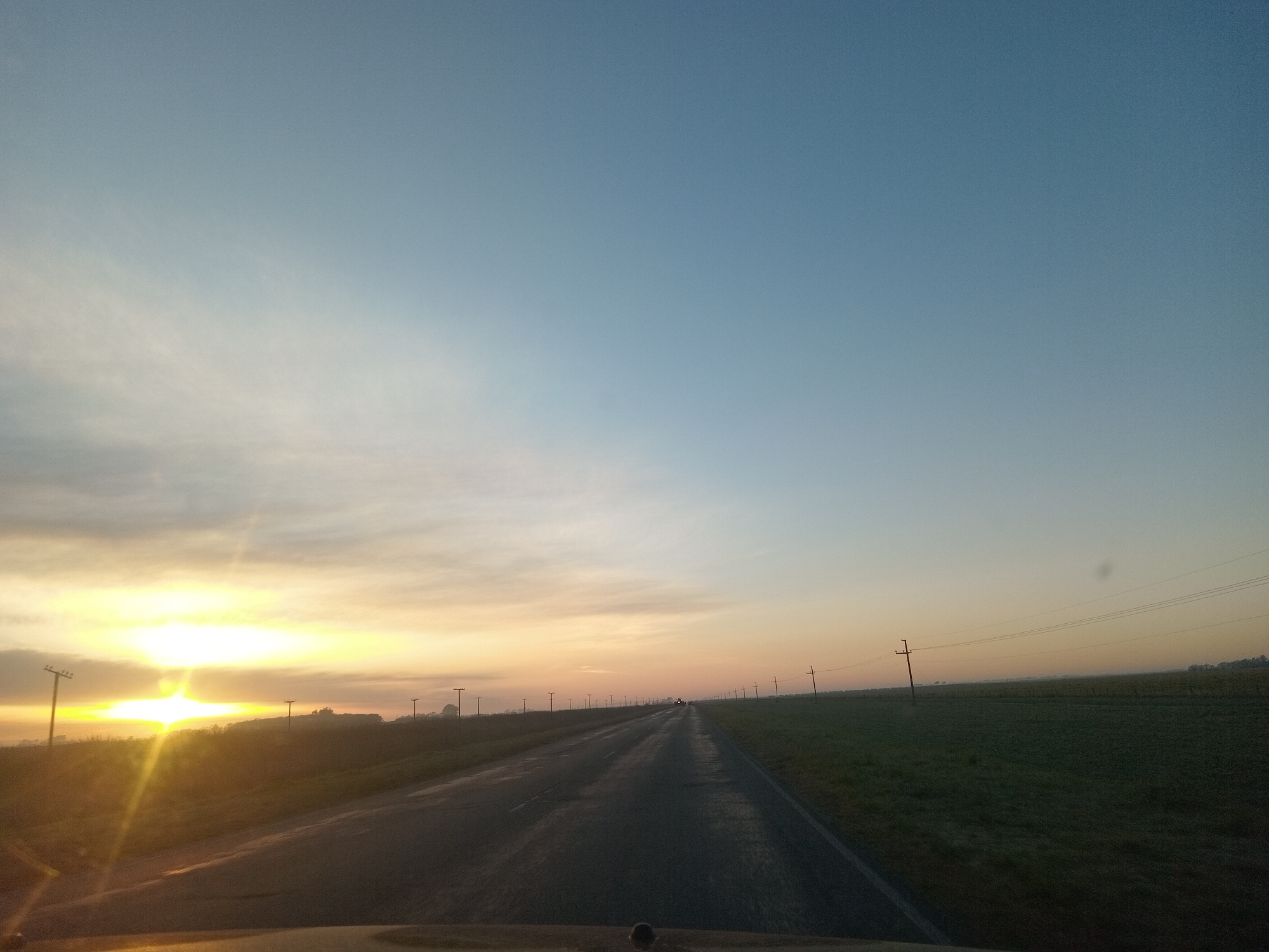 The image depicts a road stretching into the distance, flanked by fields and utility poles under a vast, clear sky. The sun, positioned on the left side of the frame, is low on the horizon, casting a bright glare and illuminating the sky with warm, orange and yellow hues. Wispy clouds are visible near the sun. The road appears to be asphalt and shows signs of wear. In the distance, several vehicles can be seen on the road. The environment suggests an open, rural setting. The perspective is from inside a vehicle, looking forward.
