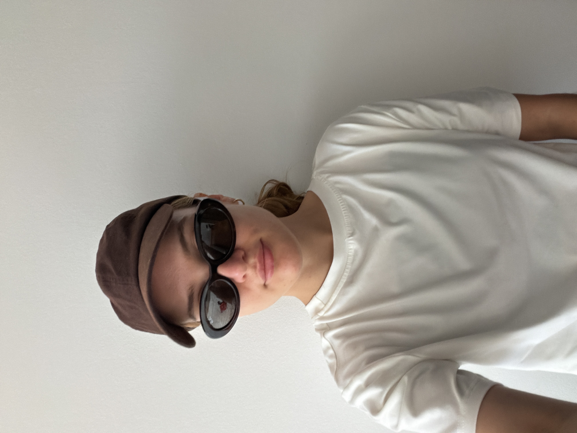 The media file is an image of a person wearing a brown cap and black sunglasses. The person is facing forward with their head tilted slightly to the side. They are wearing a white t-shirt. The background is a plain, light-colored wall. The lighting suggests it is daytime. The overall mood appears casual and relaxed. There is no visible text in the image.