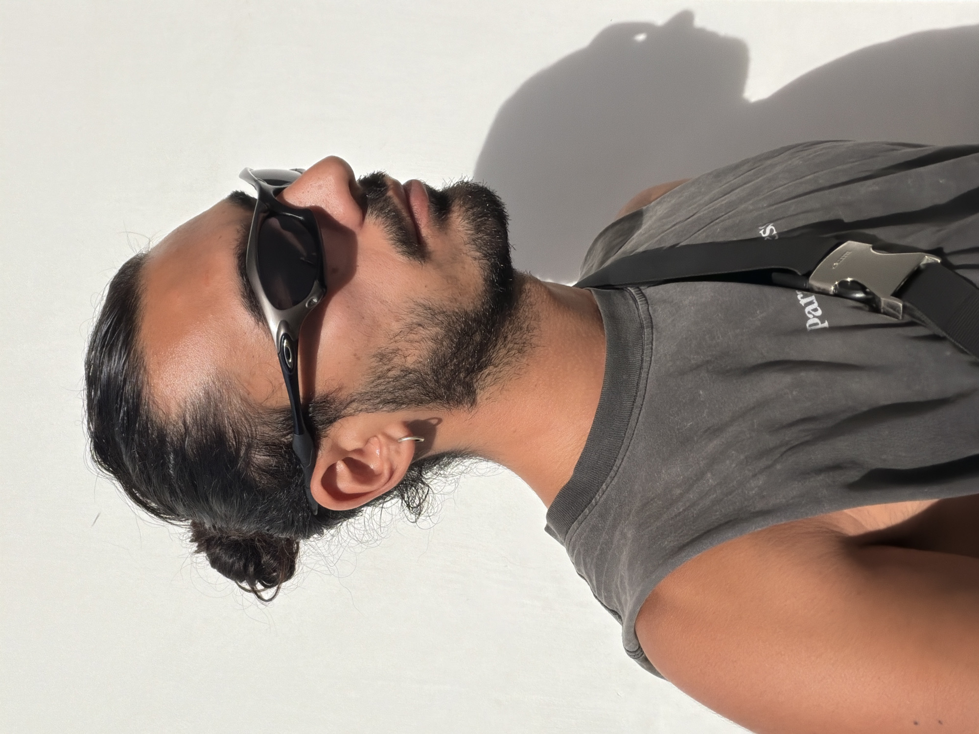 A medium shot depicts a person, likely male, with a dark beard and sunglasses, lying on a white surface in bright sunlight. The person has dark hair pulled back into a bun. They are wearing a gray sleeveless shirt with a black strap across the chest, secured with a metal buckle. The location is speculatively located in North Kuta, Indonesia.