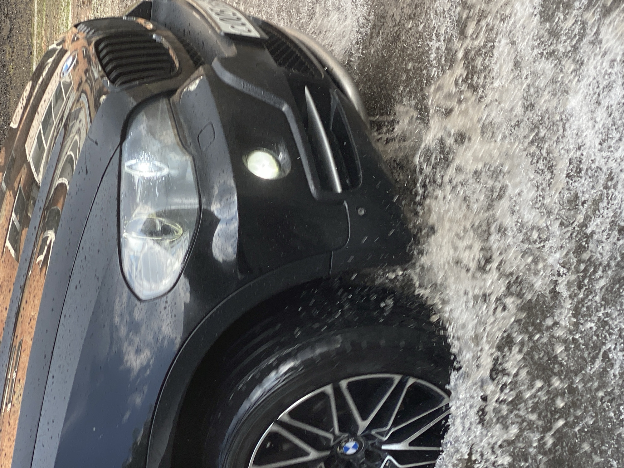 The image captures a close-up view of a black BMW SUV driving through a large puddle in Novokuznetsk, Russia. The vehicle's front wheel is splashing water as it moves. The scene appears to be on a road, likely in a residential or urban area, given the reflection of buildings on the car's hood. The lighting suggests it is daytime, possibly overcast due to the water droplets on the car and the ambient light. No people are visible in the frame. The focus is on the car and the interaction with the water.