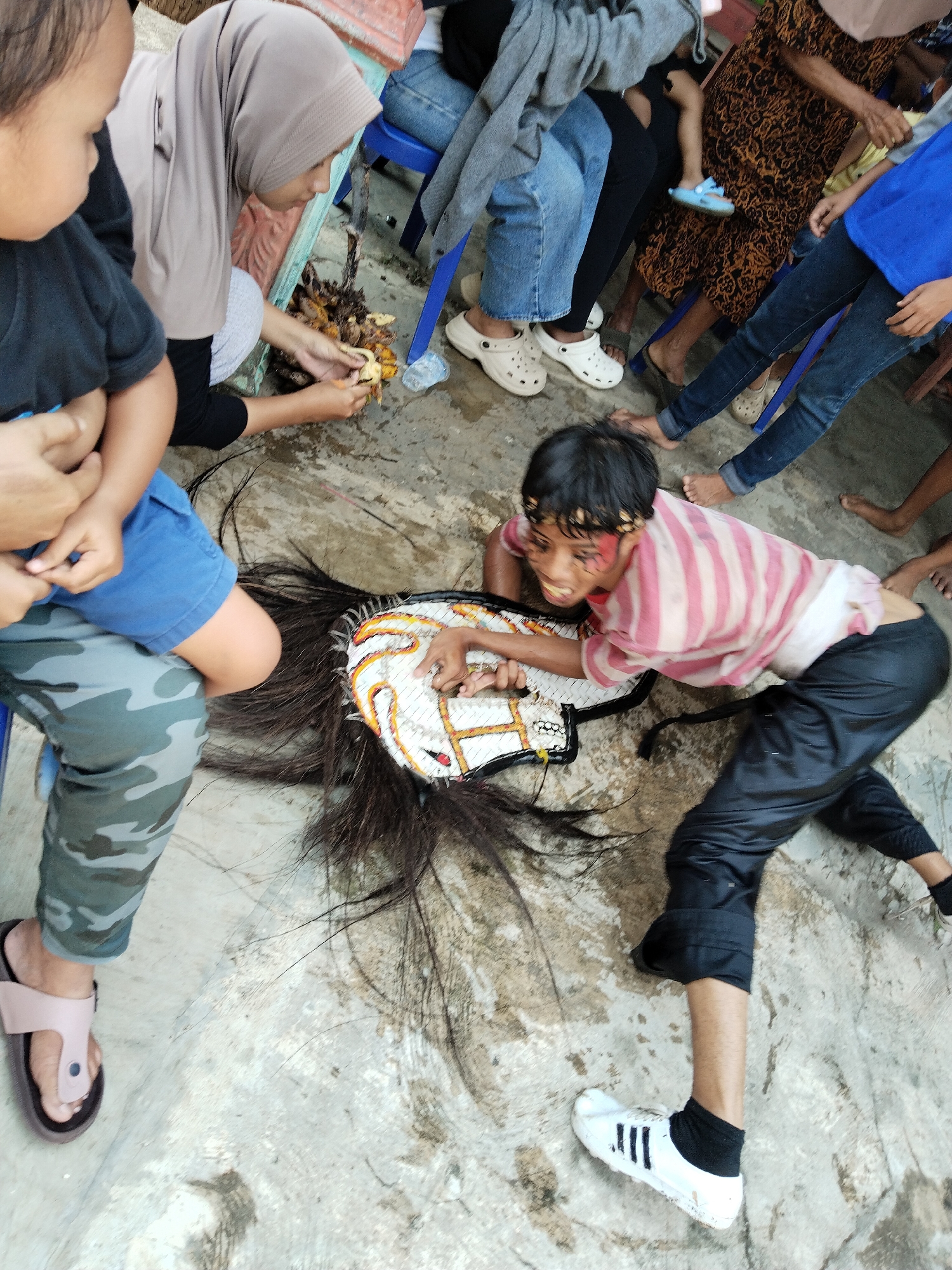 The image depicts a group of people outdoors in Pereng, Indonesia. A young boy in a pink and white striped shirt and black pants lies prone on a concrete surface, interacting with a traditional, ornate mask with attached horsehair. The boy has what appears to be painted red markings on his face. Several individuals surround him: one wearing a hijab is processing food, possibly fruit, and other people are seated or standing. Various items of clothing and footwear are visible, including jeans, sandals, sneakers, and Crocs. The environment appears to be an open, outdoor space.