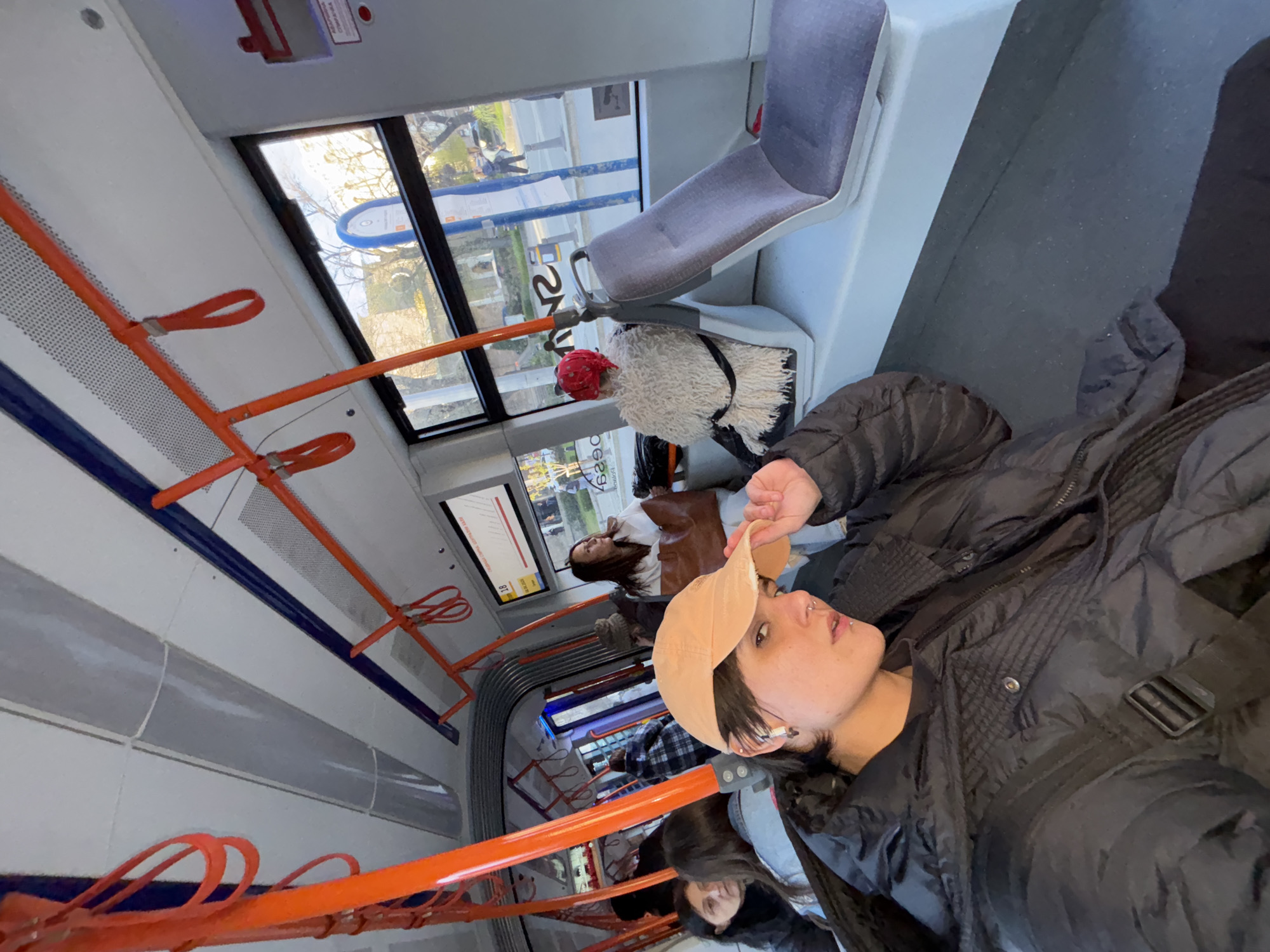 The image shows the interior of a public transport vehicle, likely a tram or bus, with several passengers. A person wearing a black jacket and a tan baseball cap is visible in the foreground. Other people are seated or standing in the background. The interior features orange handrails and grey seats. Outside the window, there is urban scenery, which, based on visual clues in the windows, is located within the city of Sofia, Bulgaria. A plush toy with a red hat is seated on a seat.
