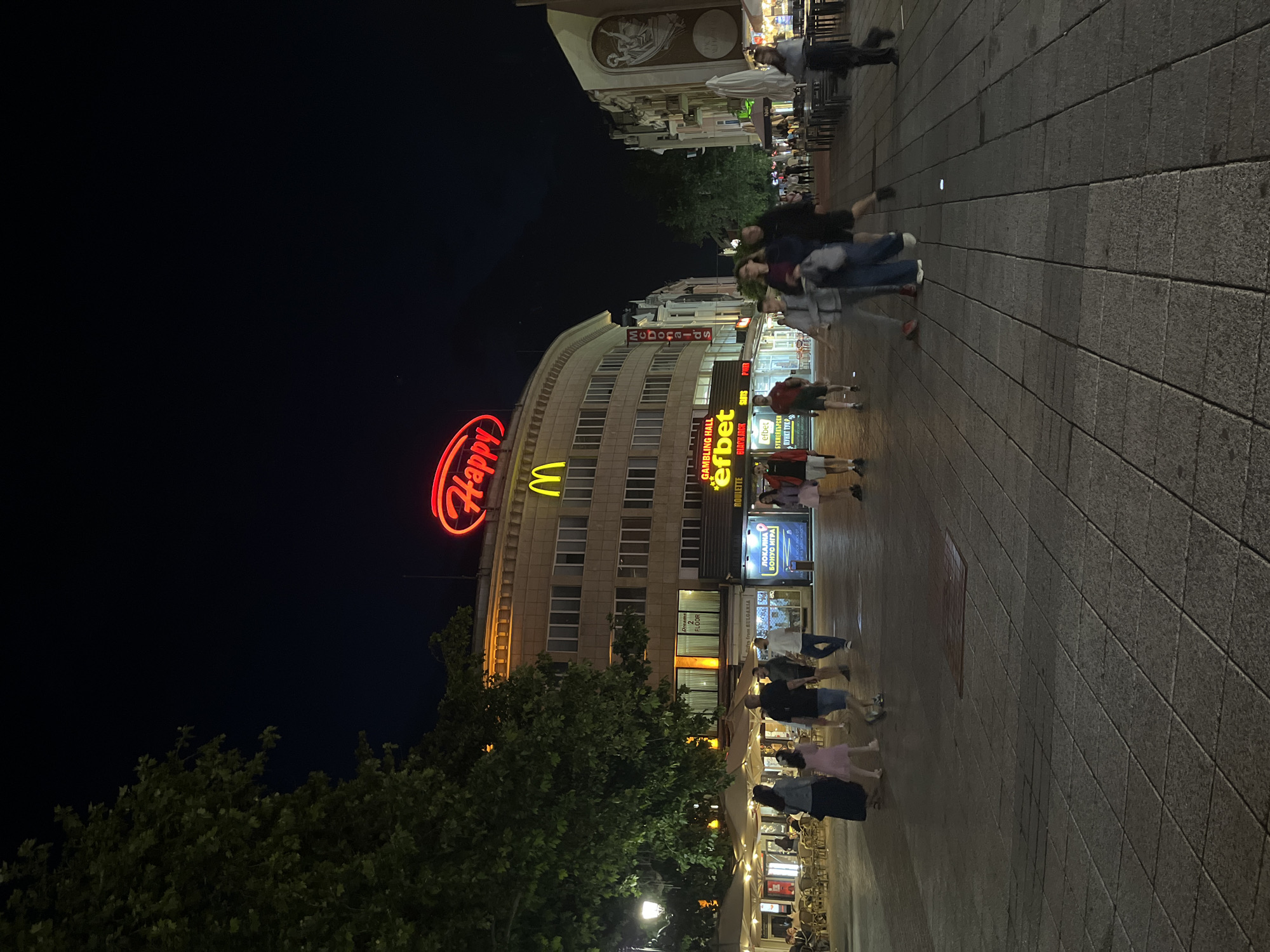 This image depicts a lively street scene at night in Plovdiv, Bulgaria. The setting is a wide, paved pedestrian thoroughfare lined with multi-story buildings, all brightly illuminated by various commercial signs and streetlights against a dark night sky.
On the left side, a prominent red neon sign boldly spells "Happy" atop a large, curved building, with the distinctive yellow "M" logo of McDonald's visible directly beneath it. Further down the street, additional signs advertise other establishments, including a "GAMBLING HALL efbet" sign promoting "ROULETTE," "BLACK JACK," and "SLOTS." Another sign displays "ЛОКАЛНА БОНУС ИГРА" (Local Bonus Game) and "БУКМЕЙКЪРСКИ ПУНКТ ТУК" (Bookmaker's Point Here). A sign on one of the buildings indicates "2nd FLOOR."
Numerous people are seen walking along the street, individually and in small groups, many appearing casually dressed. Their figures are slightly blurred due to movement and the low light, suggesting a dynamic and bustling environment. On the right side of the street, outdoor seating areas with tables and chairs are visible, implying cafes or restaurants where some people are seated. Green trees line the left foreground, adding a touch of nature to the urban setting. The overall atmosphere is vibrant and social, indicative of a city center active during the evening.