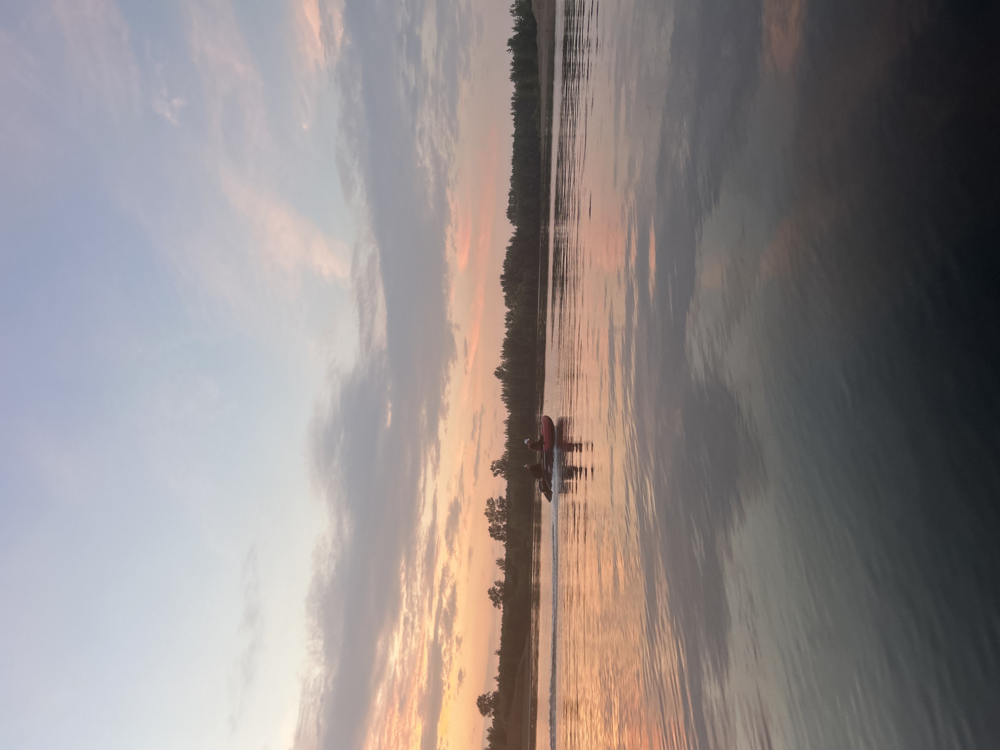 This is a horizontal, eye-level shot of a red inflatable boat with two people in it, moving across a wide, calm river at sunset. The boat is leaving a wake behind it, and its reflection is visible in the water. The sky above is a gradient of pale blue and soft pinks, with wispy clouds. The opposite bank of the river is lined with a dense, dark green forest, and the sky is reflected in the water, creating a mirror image of the sunset colors. The scene suggests a peaceful evening on the river in Noviy, Russia.