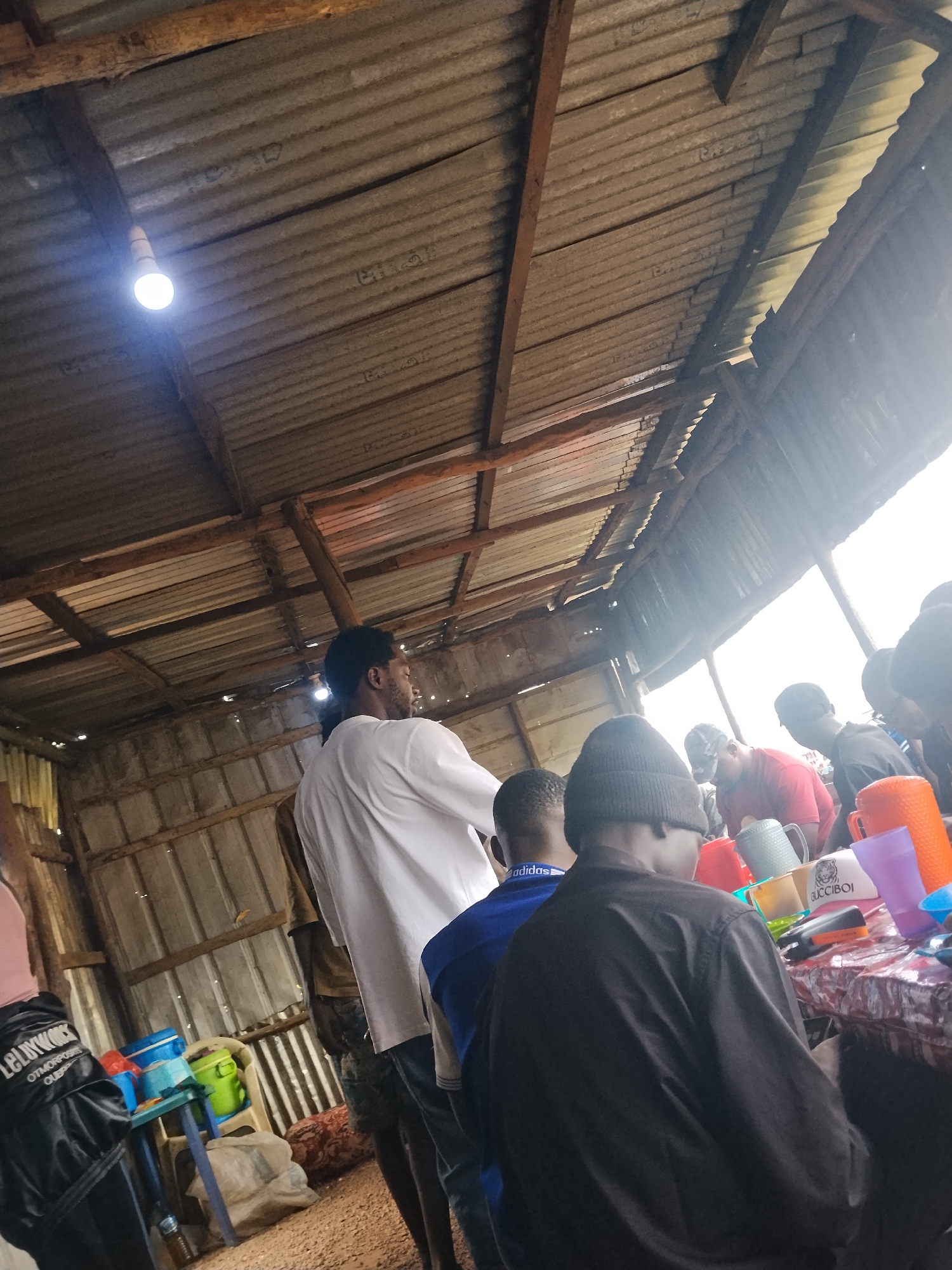 In Durumi, Nigeria, this image depicts an interior scene of a shaded open-air market stall. A corrugated metal roof, supported by wooden beams, covers the area. The light from a single bulb illuminates the space, casting shadows and highlighting the weathered textures of the corrugated metal and wooden supports.
Several people are present, mostly seen from the back or side. One man, facing away from the camera, wears a white long-sleeved shirt. Others are seated or standing around a table laden with colorful plastic cups and mugs. The items on the table include orange, purple, and blue cups, as well as what appear to be metal mugs. A black jacket with white lettering is visible on the left, and other individuals are wearing casual attire, including a black beanie and a blue shirt with an Adidas logo. The ground appears to be dirt or gravel. The overall atmosphere suggests a casual gathering or a vendor's stall at a market.