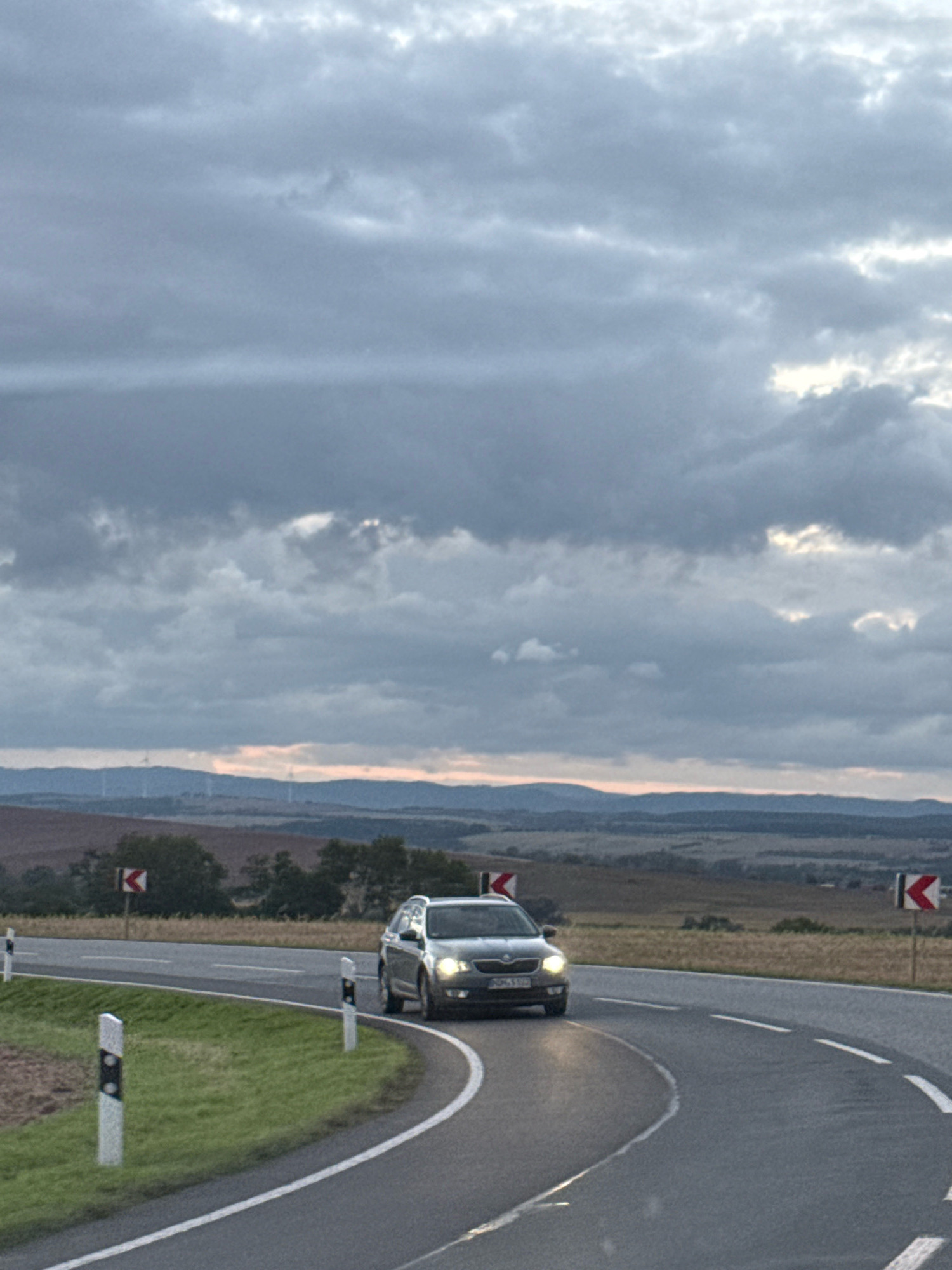 A dark-colored Skoda Octavia estate car is driving on a winding asphalt road with white lane markings. The car is in the foreground, heading towards the right side of the frame. The road is bordered by a grassy verge on the left, marked by bollards with reflective white and black stripes. On the right side of the road, there are traffic signs indicating curves. In the background, a hilly landscape unfolds under a cloudy sky. The sky is filled with a mix of gray and white clouds, with some hints of light breaking through. In the distance, wind turbines can be seen on the hills. The scene appears to be an outdoor landscape, possibly in a rural area outside of Nordhausen, Germany, given the presence of wind turbines and rolling hills. The lighting suggests it is either early morning or late afternoon.