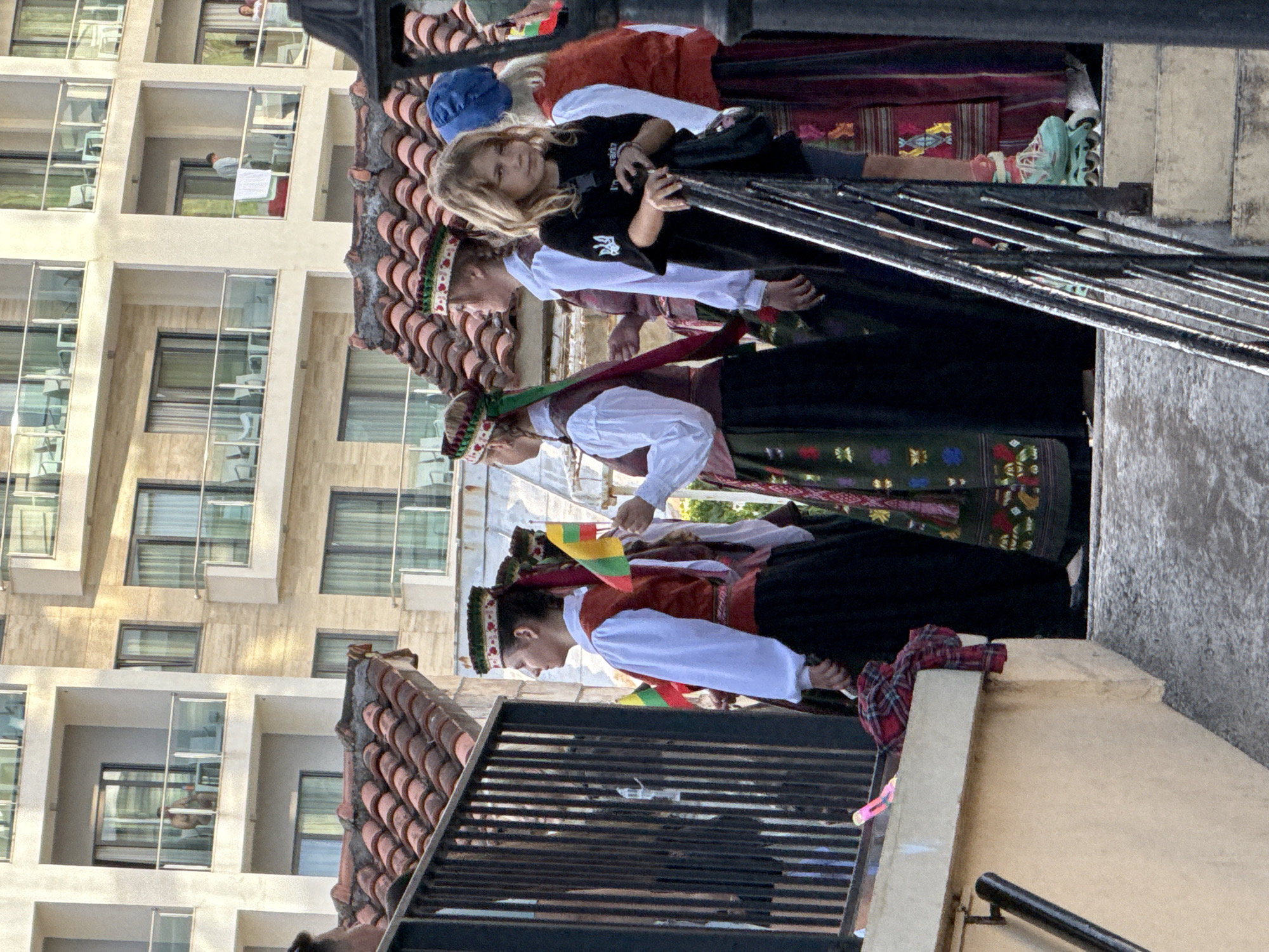 The image depicts an outdoor daytime scene in Budva, Montenegro. In the foreground and midground, a group of individuals in traditional folk costumes stands on a slightly elevated area, while a child in modern attire stands on concrete steps below them. A large, multi-story building with numerous balconies dominates the background.
The group in traditional dress consists of at least four adults, predominantly female, wearing white long-sleeved blouses, dark vests (some reddish-brown, others darker), and long, dark skirts with embroidered patterns. Their heads are adorned with elaborate, colorful headpieces featuring floral or geometric designs and ribbons (green and maroon ribbons are visible). Two individuals within this group are holding small flags with horizontal stripes of yellow, green, and red, which are the colors of the Lithuanian flag.
To the right, a young blonde-haired girl, appearing to be pre-teen, is prominently featured. She wears a black short-sleeved t-shirt with a white graphic on the left sleeve (a trident-like symbol) and white text on the chest. She has grey shorts, pink socks, and light blue roller skates. She holds onto a dark metal railing with both hands and looks directly towards the viewer. A small black shoulder bag is slung across her body.
The immediate environment includes a dark metal railing on the right, leading down concrete steps where the child is positioned. To the left, a beige concrete ledge holds a red and black plaid cloth and a pink plastic object. Behind the foreground elements and the traditionally dressed group, a red-brown clay tiled roof is visible.
In the background, a modern, light-colored (beige/cream) multi-story building with a flat roof features multiple balconies with glass railings. Each balcony contains light-colored outdoor furniture. Several individuals are visible on these balconies: one shirtless male on an upper-left balcony stands by a table with a drink; another individual in a light-colored shirt on a middle-right balcony drapes a white towel over the railing; and a third person in a white garment is partially visible on an adjacent balcony to the right. The building's windows are dark-framed sliding glass doors. The sky is bright and clear.
