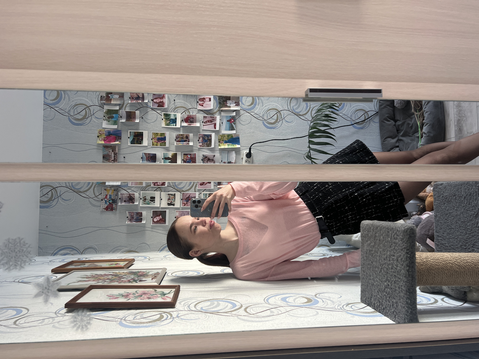 A mirror reflects a woman taking a selfie while lying on a bed or floor in a room, which is adorned with floral-patterned wallpaper and photographs tacked to the wall. The woman is wearing a pink long-sleeved top and a black patterned skirt with a black belt. Three framed pieces of artwork are visible on the surface near her. A cat tree stands near the woman's feet. It is unknown if the location is Chaikovsky, Russia.