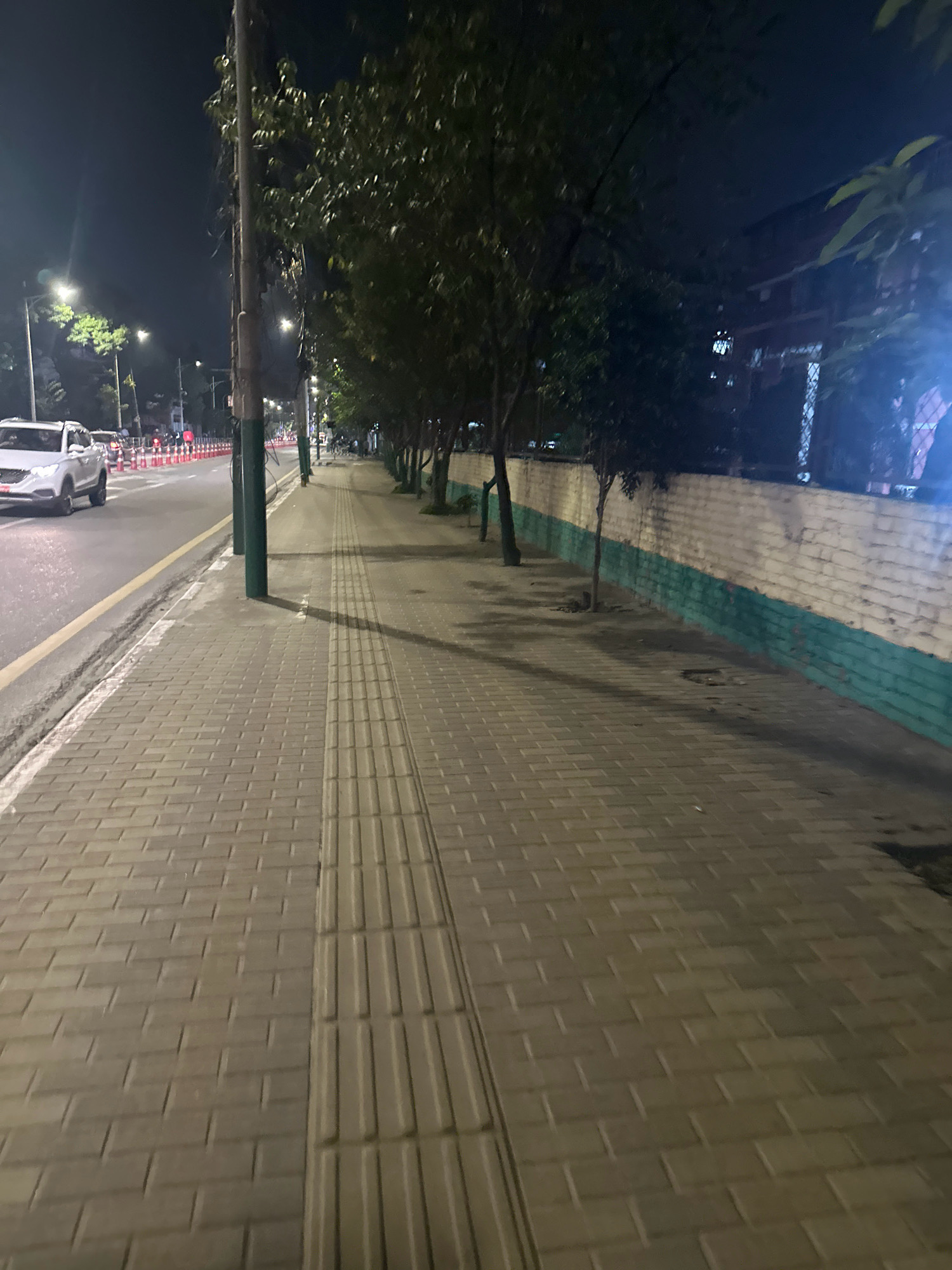 A pedestrian sidewalk made of rectangular pavers runs parallel to a street at night. The sidewalk has a tactile paving strip for the visually impaired along its edge. The street is illuminated by streetlights and car headlights. A white SUV is visible driving on the street. Trees line the sidewalk, and a white brick wall with a green band at the bottom runs behind them. The sky is dark, indicating nighttime. The scene appears to be a typical urban street in Kathmandu Metropolitan City, Nepal, at night.