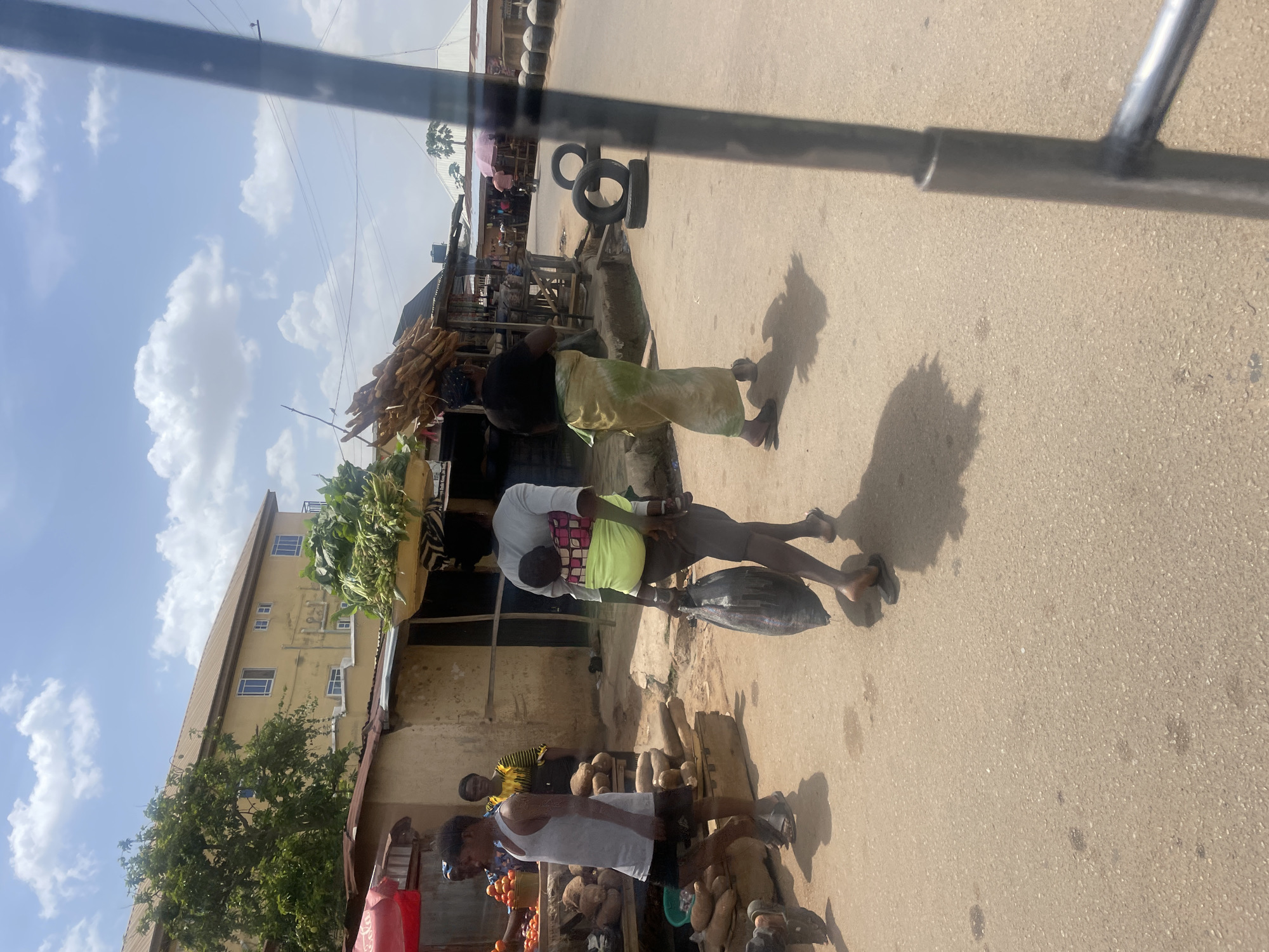 The image depicts an active outdoor market scene on a sunny day in what appears to be a local community, consistent with Jos, Nigeria. The photograph is taken from a slightly elevated perspective, possibly from a vehicle, with a black metal bar crossing the upper portion of the frame.
Several people are visible engaging in market activities. In the foreground, a person wearing a white tank top and dark shorts stands near a stall displaying large root vegetables, likely yams, and some red fruit, possibly tomatoes. Next to them, another individual in a yellow and black striped shirt observes the scene.
Further back, a person in a white top and green waist wrap is seen carrying a large, dark-colored fish by its tail, walking away from the camera. Behind them, another individual, dressed in a black top and a light-colored, possibly patterned skirt or wrap, walks with a load on their head, which appears to be firewood or stacked corn cobs. Both of these individuals are wearing flip-flops, and their shadows are distinctly cast on the paved ground, indicating bright midday sun.
The market features various makeshift stalls constructed from wood and corrugated metal, displaying produce. A tall, light yellow multi-story building with multiple windows stands in the background on the left. The ground is a paved road, and several black car tires are placed along the roadside in the mid-ground, possibly as barriers or for sale. The sky is bright blue with scattered white clouds, and power lines are visible overhead. No discernible text is visible in the image.