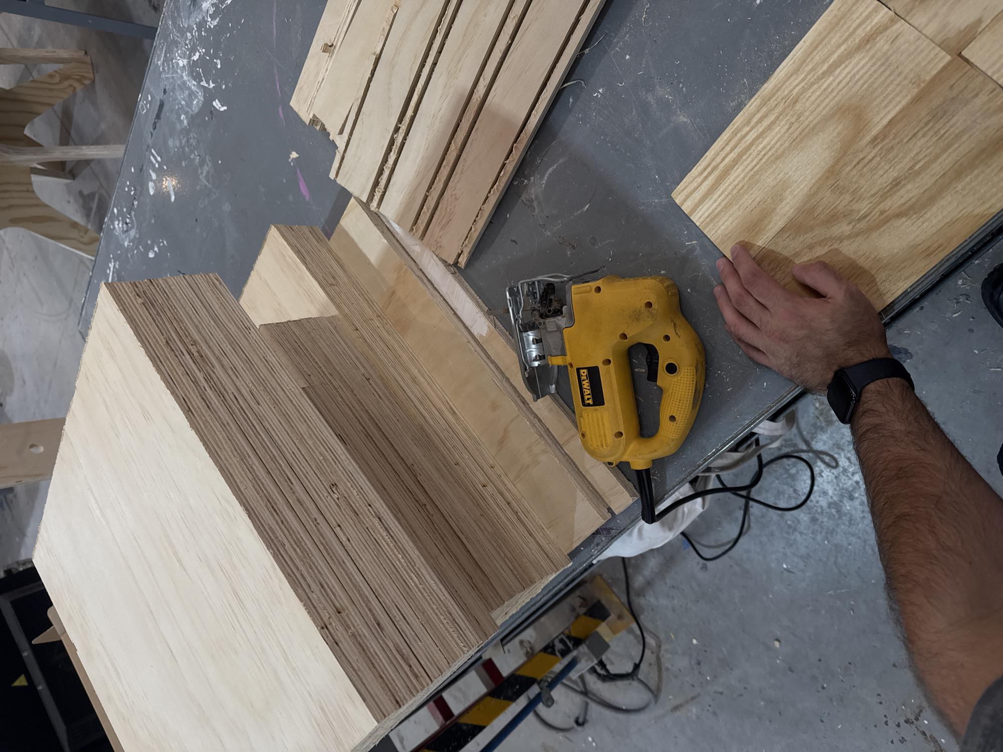 This image provides an overhead view of a busy woodworking or crafting area, likely a workshop, in San Andrés Cholula, Mexico. The scene is dominated by wood materials and tools, indicating an ongoing fabrication project.
In the foreground, a person's right hand and forearm are visible. The arm is hairy, and a black smartwatch is worn on the wrist. The hand is gently placed on a rectangular piece of light-colored wood, suggesting manipulation or preparation. Adjacent to the hand and slightly to the left, a yellow and black DeWalt jigsaw rests on a gray workbench. Black electrical cords snake across the surface, leading away from the tool. The workbench itself is mottled with white and purple paint splatters, signs of heavy use and creative activity.
Several stacks of plywood or laminated wood panels are arranged across the workspace. A substantial, thick stack stands vertically on the left, while thinner, horizontally stacked panels are positioned centrally. Near the top right, additional long, narrow cut pieces of wood are neatly arranged. The abundance of raw and cut wood, along with the presence of the jigsaw, strongly suggests that the primary activity involves cutting, shaping, or assembling wood.
In the blurred background, other wooden structures, possibly unfinished projects or storage, can be discerned. A distinct black and yellow striped warning tape is visible on a lower structure, reinforcing the industrial or workshop setting. The lighting suggests daytime, likely indoors with artificial illumination. The only visible text, "DEWALT," clearly labels the jigsaw. The overall impression is one of practical, hands-on work in a dedicated creative or industrial environment.