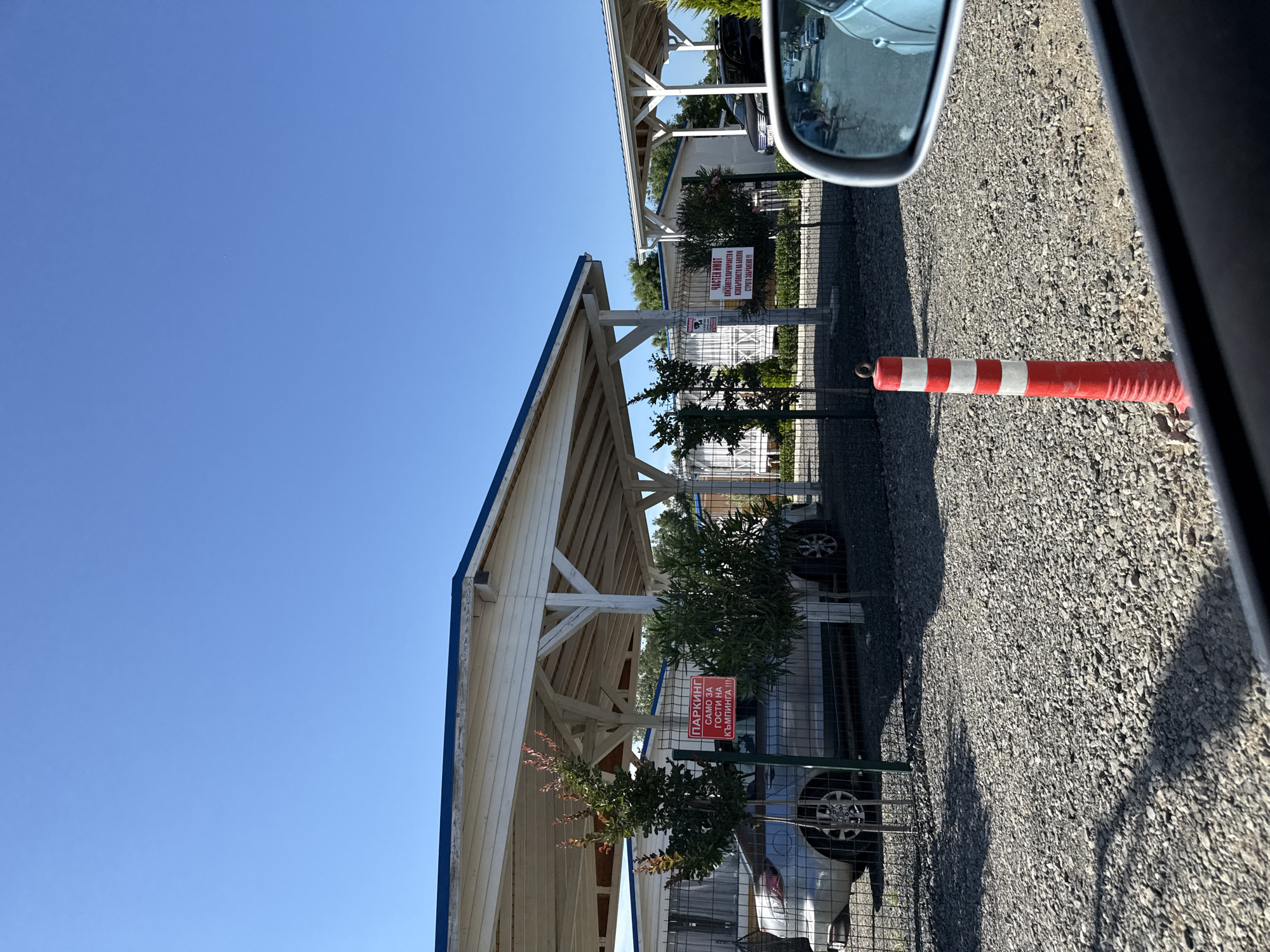 The image shows a sunny day in Tsarevo, Bulgaria, with a clear blue sky. In the foreground, a gravel parking area is visible. A red and white striped traffic cone stands near the center. A car's side mirror reflects a street with parked cars. Beyond the cone, a fence encloses a car park with covered parking structures. Two signs are mounted on the fence. The sign on the left is red and reads "ПАРКИНГ САМО ЗА ГОСТИ НА КЪМПИНГА!!!" (Parking only for campsite guests!!!). The sign on the right is also red and reads "ЧАСТЕН ИМОТ ДИ ЗАВЕТЯ ЗАБРАНЕНО И СТРОГО ЗАБРАНЕНО!!!" (Private property. Entry forbidden and strictly forbidden!!!). A silver car is parked under one of the covered structures. Two small trees are planted in front of the fence. In the background, more covered parking structures and buildings are visible.