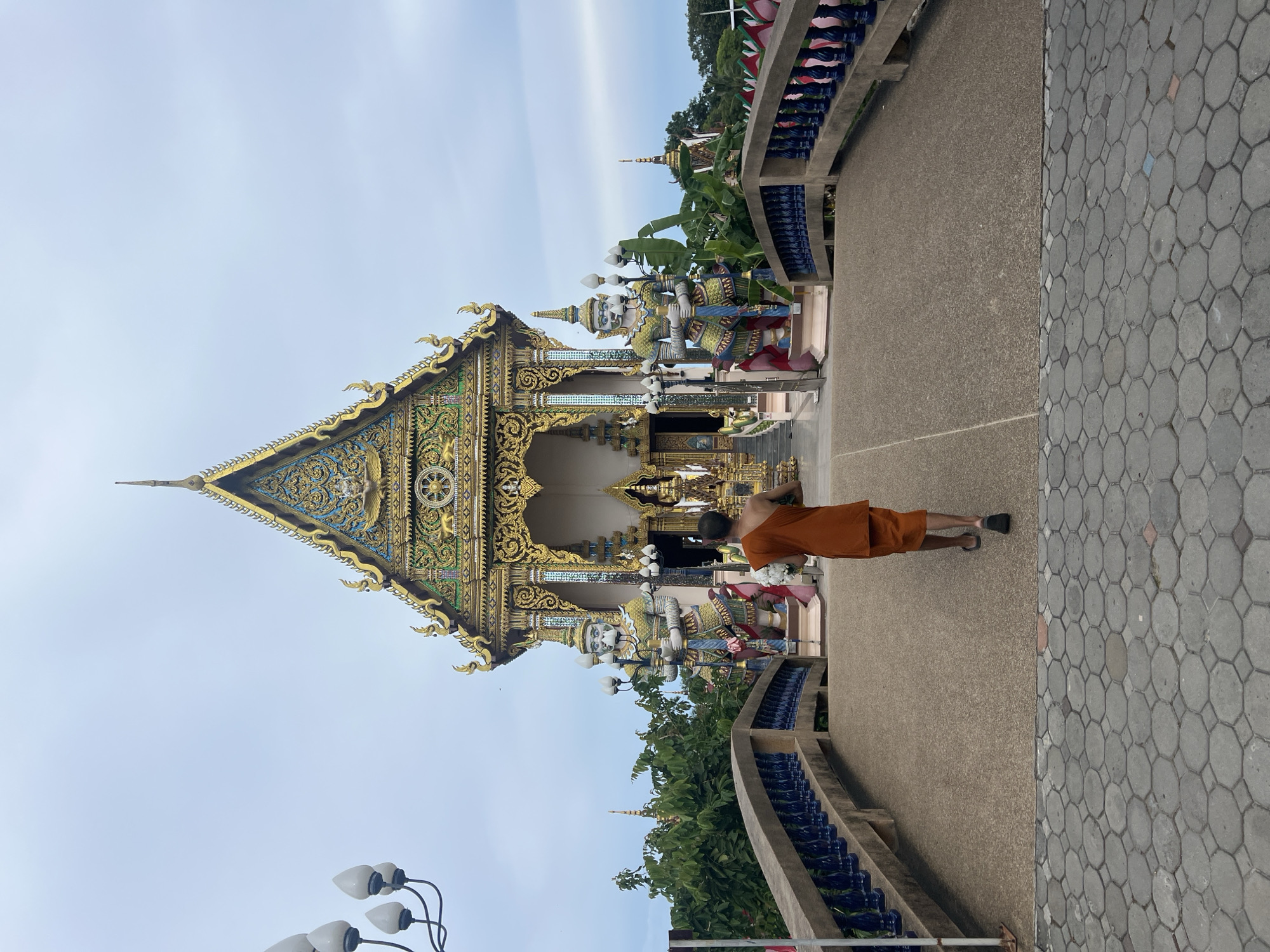 A Buddhist monk walks across a bridge towards a temple in Baan Plai Laem, Thailand. The monk, wearing an orange robe, carries a bundle of white flowers. Two large guardian statues stand on either side of the temple entrance. The temple is ornate with gold and colorful decorations. The bridge has blue railings and a concrete walkway. The ground is paved with hexagonal gray stones. The sky is overcast.