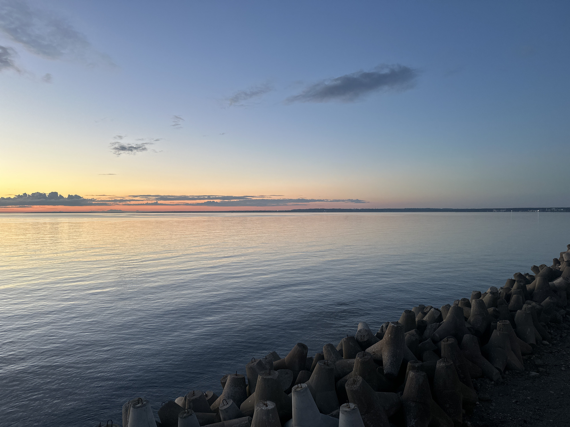 The image depicts a serene twilight scene at the Tallinn coast. In the foreground, a dense arrangement of tetrapods, a type of concrete coastal defense, lines the shoreline, creating a textured barrier. These structures are dark gray and appear weathered. Beyond the tetrapods, a calm body of water stretches out, reflecting the soft hues of the sky. The water's surface exhibits subtle ripples, suggesting a gentle breeze. In the distance, a faint, low-lying coastline is visible, dotted with distant lights, hinting at urban development. The sky above transitions from a pale yellow and orange near the horizon, indicating the setting or rising sun, to a soft blue higher up. A few scattered clouds, wispy and dark, drift across the upper portion of the sky. The overall impression is one of tranquility and natural beauty at dusk.
