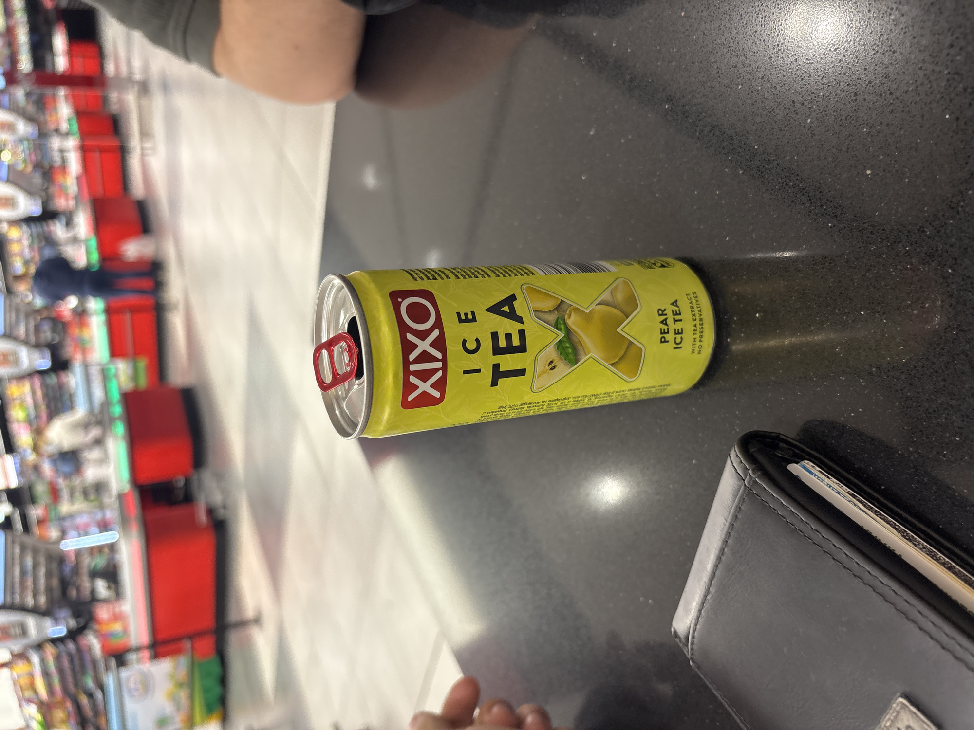 The image shows a can of Xixo Pear Ice Tea on a black, speckled countertop next to a wallet. The can is yellow with red branding and has been opened. In the blurry background, there is a section of a shop with red shelving. The image was likely taken in Sofia, Bulgaria based on the Xixo brand being available in that country.