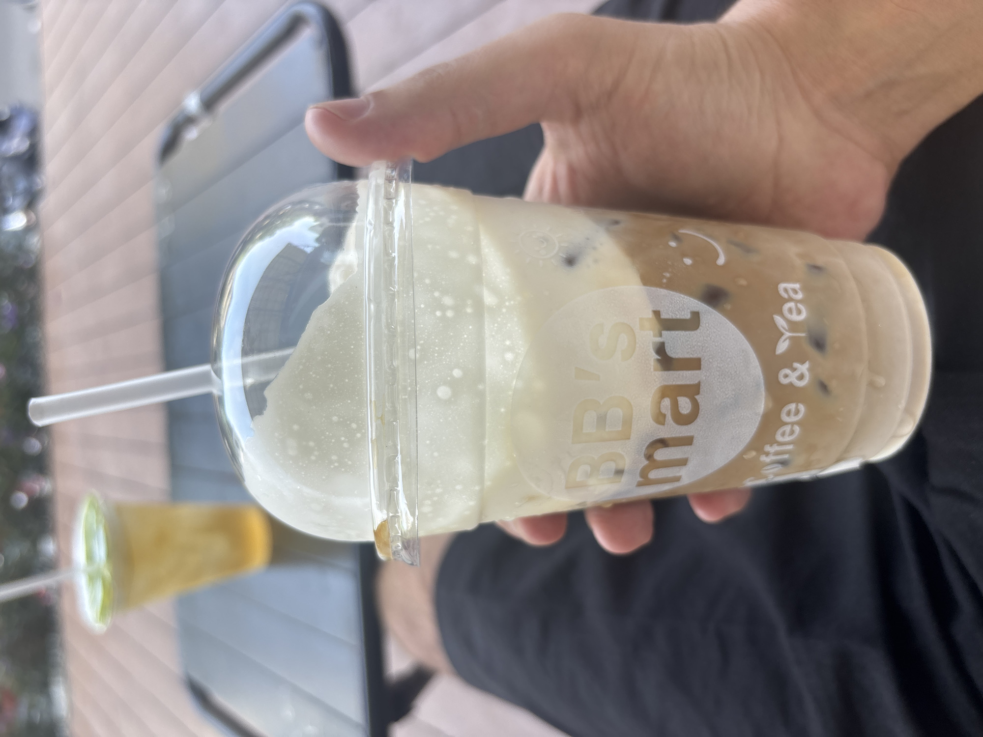 A hand holds a clear plastic cup with a domed lid, containing a layered beverage. The drink appears to be iced coffee with a frothy, cream-colored topping. The cup has a logo with the text "BB's mart" and "Coffee & Tea" printed on it. In the background, another iced drink in a clear plastic cup with a green top and a straw is visible, sitting on a dark surface. The environment suggests an outdoor seating area, possibly a cafe or restaurant, with a wooden deck-like flooring and some blurred greenery in the distance.