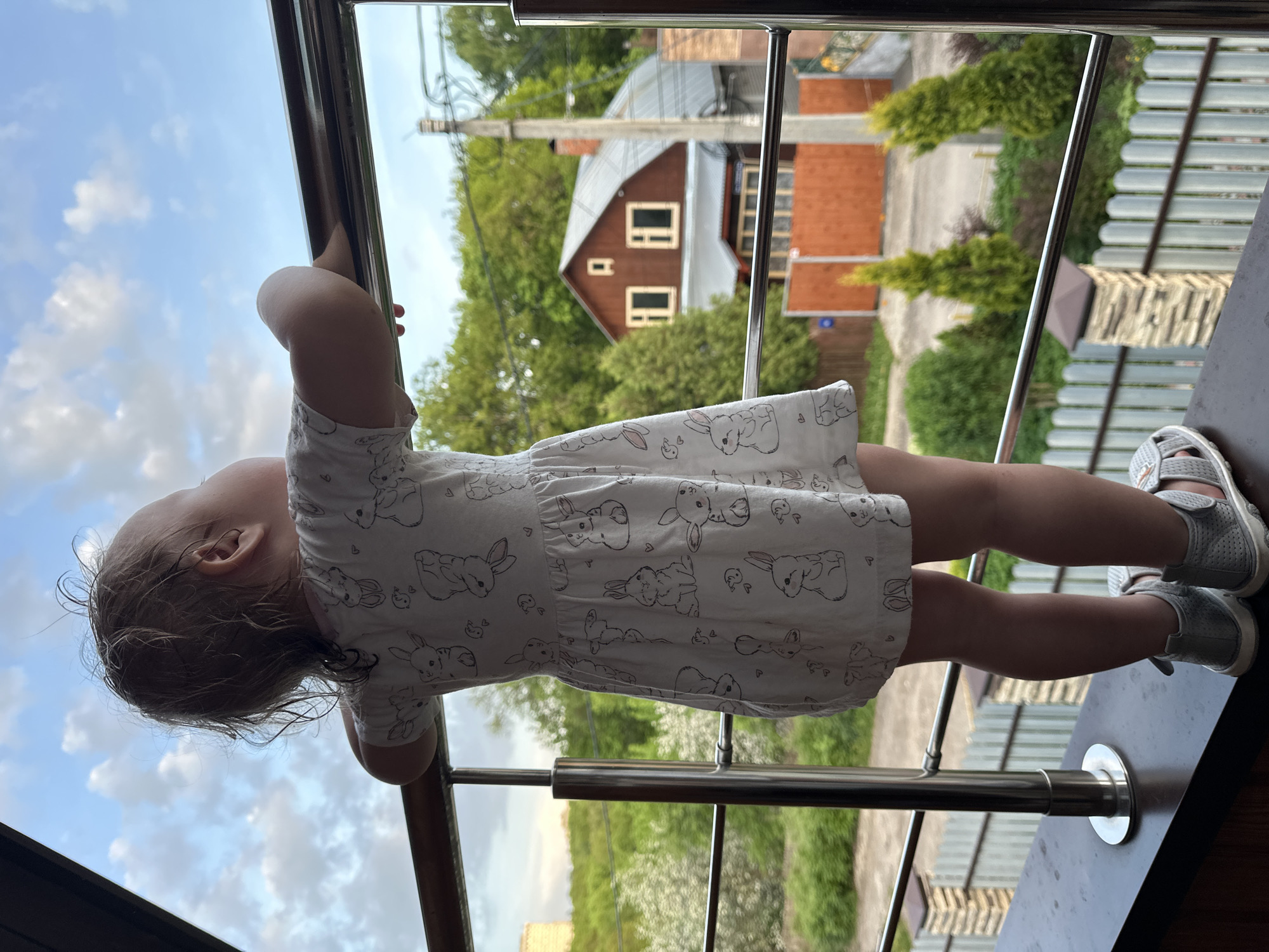 A young child, likely female, stands on a balcony. The child has fair skin and light brown hair, wearing a white dress patterned with bunny designs and grey shoes. The child grips the railing of the balcony, looking out towards the surroundings.
The balcony has a dark, possibly wooden base, and a metal railing. The view from the balcony includes greenery, a two-story house with a brown exterior and a grey roof, and a partial view of the sky with clouds. The house design and the presence of other visible structures suggest this is a residential area. The location is indicated as being in Nizhny Novgorod, Russia, based on additional context.