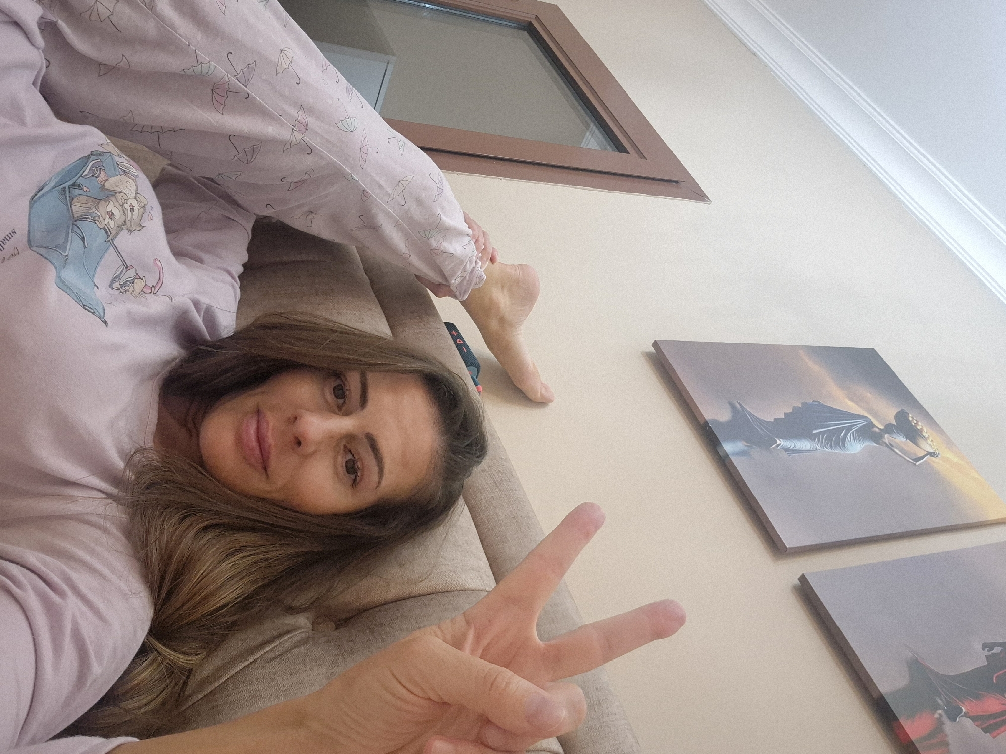 A woman in light purple pajamas lies on a beige couch, holding up a hand in a peace sign. She is positioned at an angle to the camera, with her head near the bottom left corner and her feet angled towards a brown-framed window in the top center. The pajamas have a print of umbrellas and a cartoon figure. Two square artworks adorn the beige wall next to the couch, featuring images with a sky and figures. The ceiling is white with a decorative molding. The location is unknown.