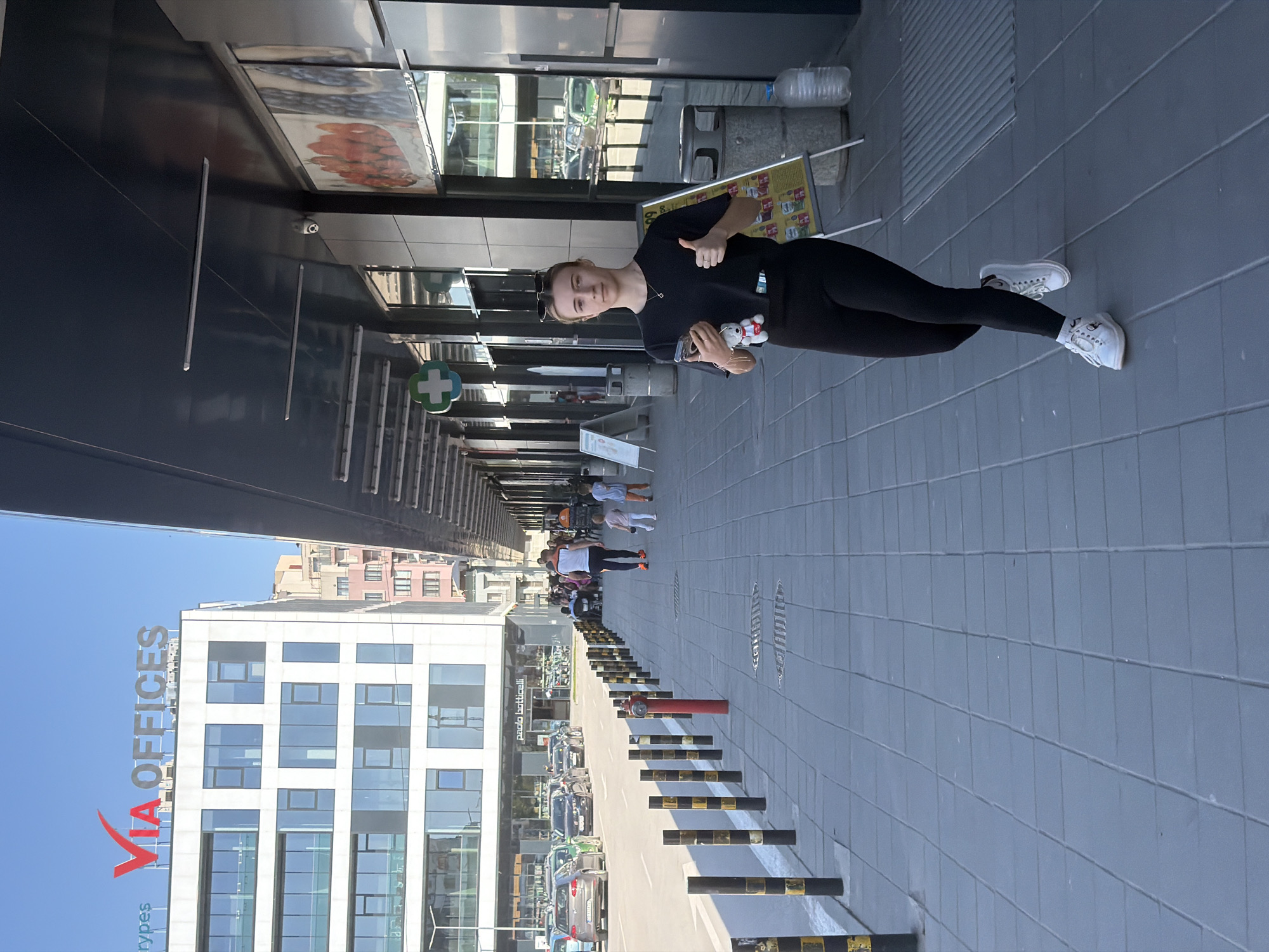 A young woman is posing for a photo in Plovdiv, Bulgaria, in front of the Via Offices building. She is standing on a tiled sidewalk, giving a thumbs-up, holding a mobile phone and a small white toy. She is wearing a black t-shirt, black leggings, and white sneakers. Behind her is the Via Offices building with its distinctive red logo, a row of black and yellow bollards, parked cars, and pedestrians walking further down the street. Above the woman is the overhang of a building, reflecting a partial view of the street. A green and white pharmacy sign is visible on the overhang, along with signage for Paolo Botticelli. The sky is blue and clear.