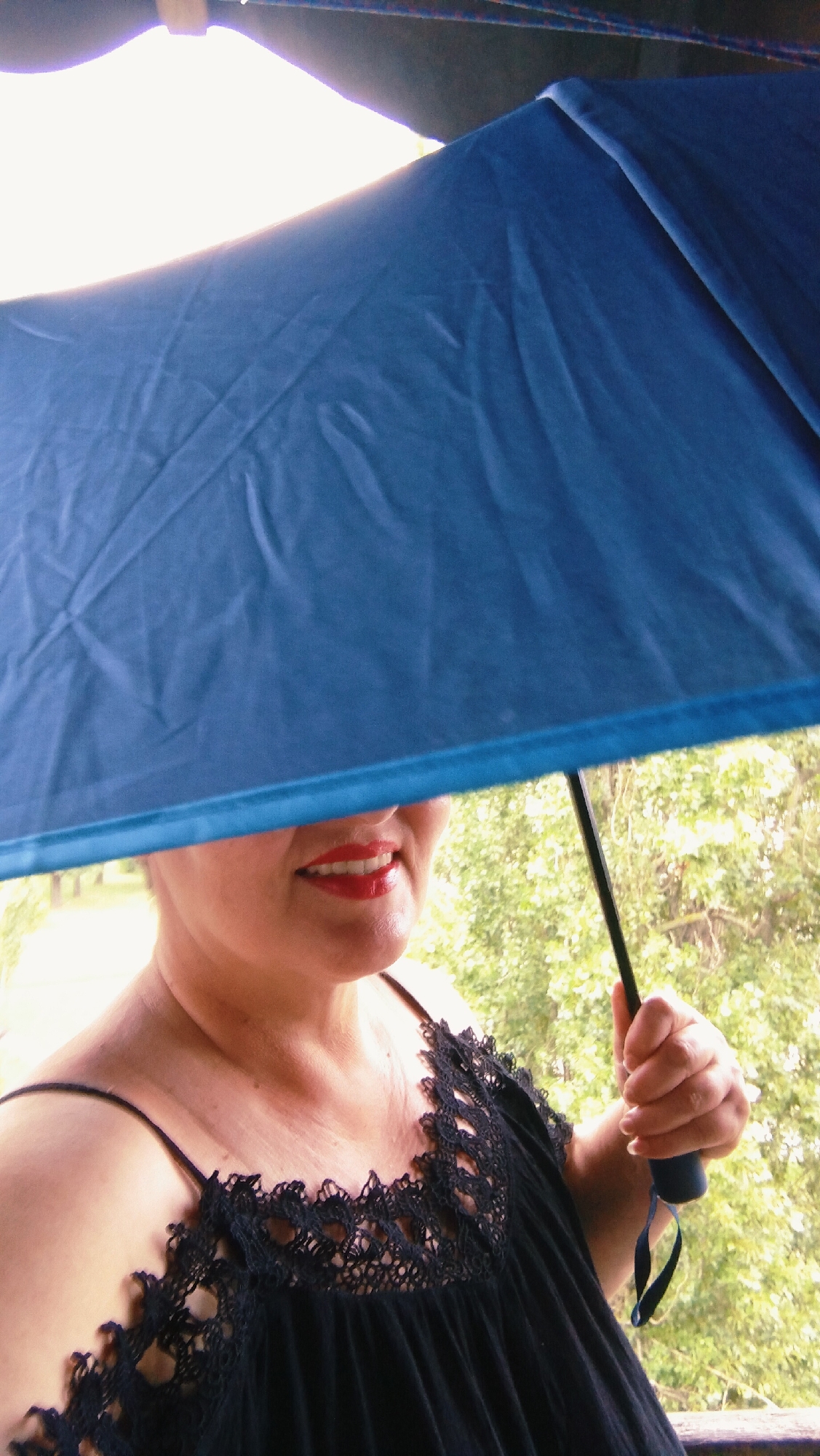 A woman is holding a blue umbrella over her head and a portion of her face. She is wearing a black top with lace trim. The background shows greenery and what appears to be a balcony or outdoor railing. The lighting suggests an overcast day. The scene is likely outdoors in a residential or park area.