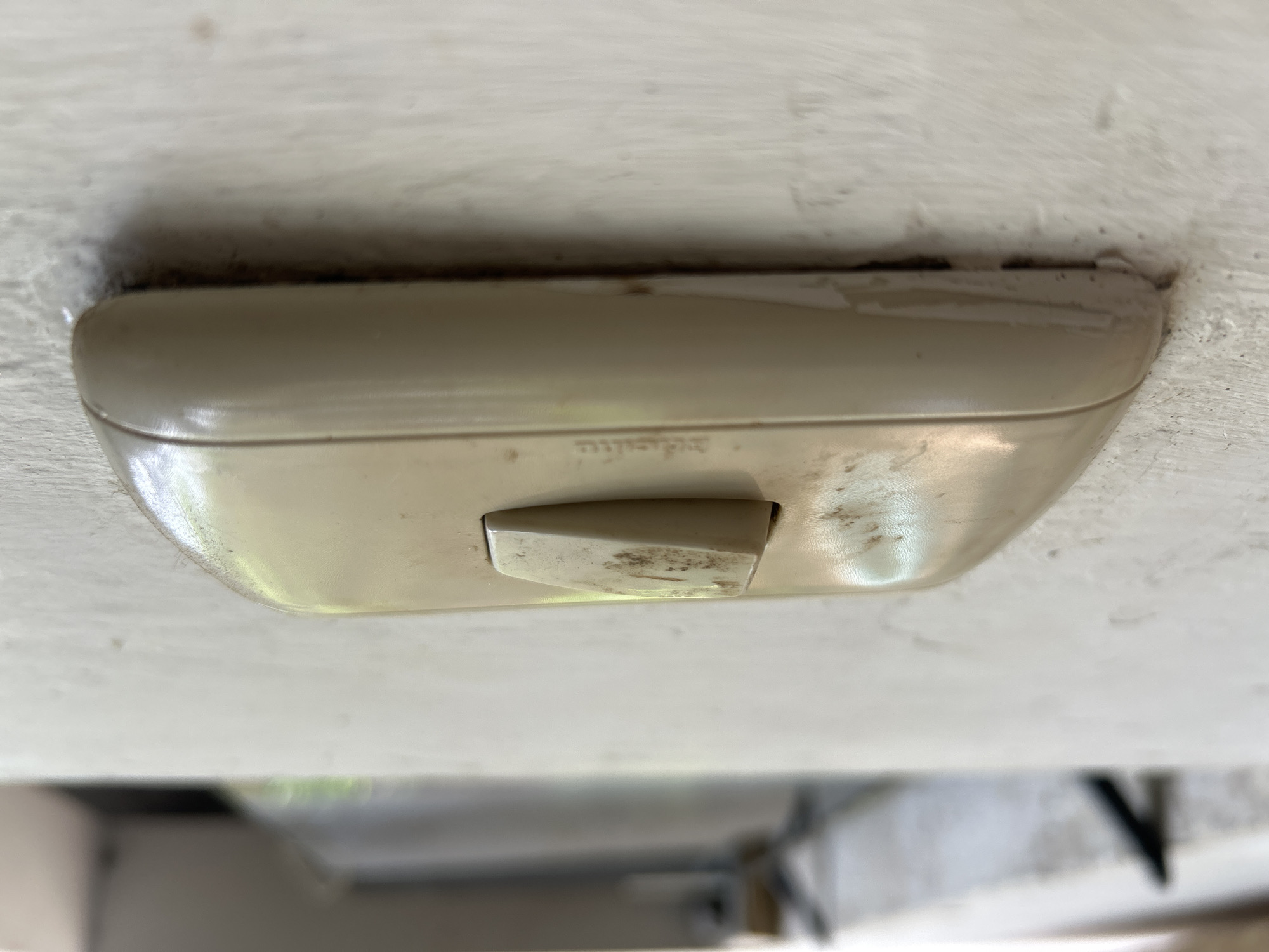 A close-up, side-view, eye-level photograph shows a beige, slightly curved light switch with a prominent beige toggle protruding from its center. The toggle is positioned in the "off" position and is visibly stained with dark, gritty marks, suggesting dirt or grime. The light switch plate is also marked with similar blemishes and shows some signs of age or wear, with a slightly discolored surface. The switch is mounted on a textured, off-white wall that appears to be stucco or a similar material. The background is out of focus, showing a blurry, light-colored area that could be a window frame or doorway, with hints of green and gray from the environment beyond. The overall impression is one of a somewhat aged and used electrical fixture in a home or building. The specific location in Santa Ana, Venezuela, cannot be determined from the visual information alone.