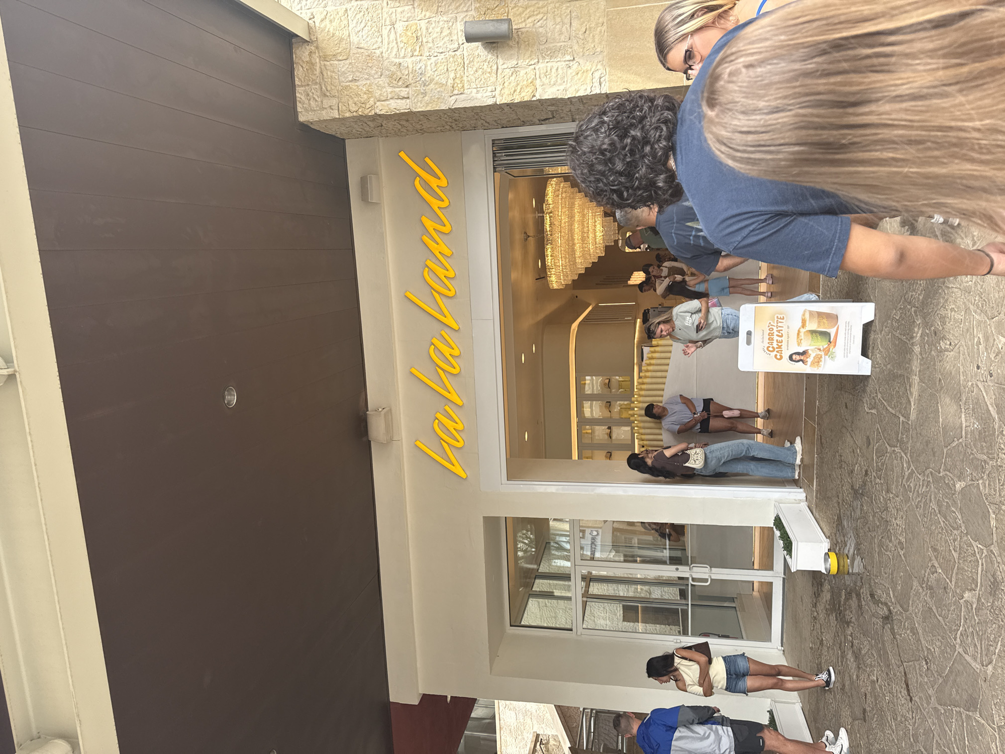 The image shows the exterior of a store called "La La Land" in San Antonio, Texas. People are waiting in line outside the store. There is a sign advertising "Carrot Cake Latte." The exterior of the building is a combination of stone and brown wood siding. A large chandelier is visible through the entrance.
