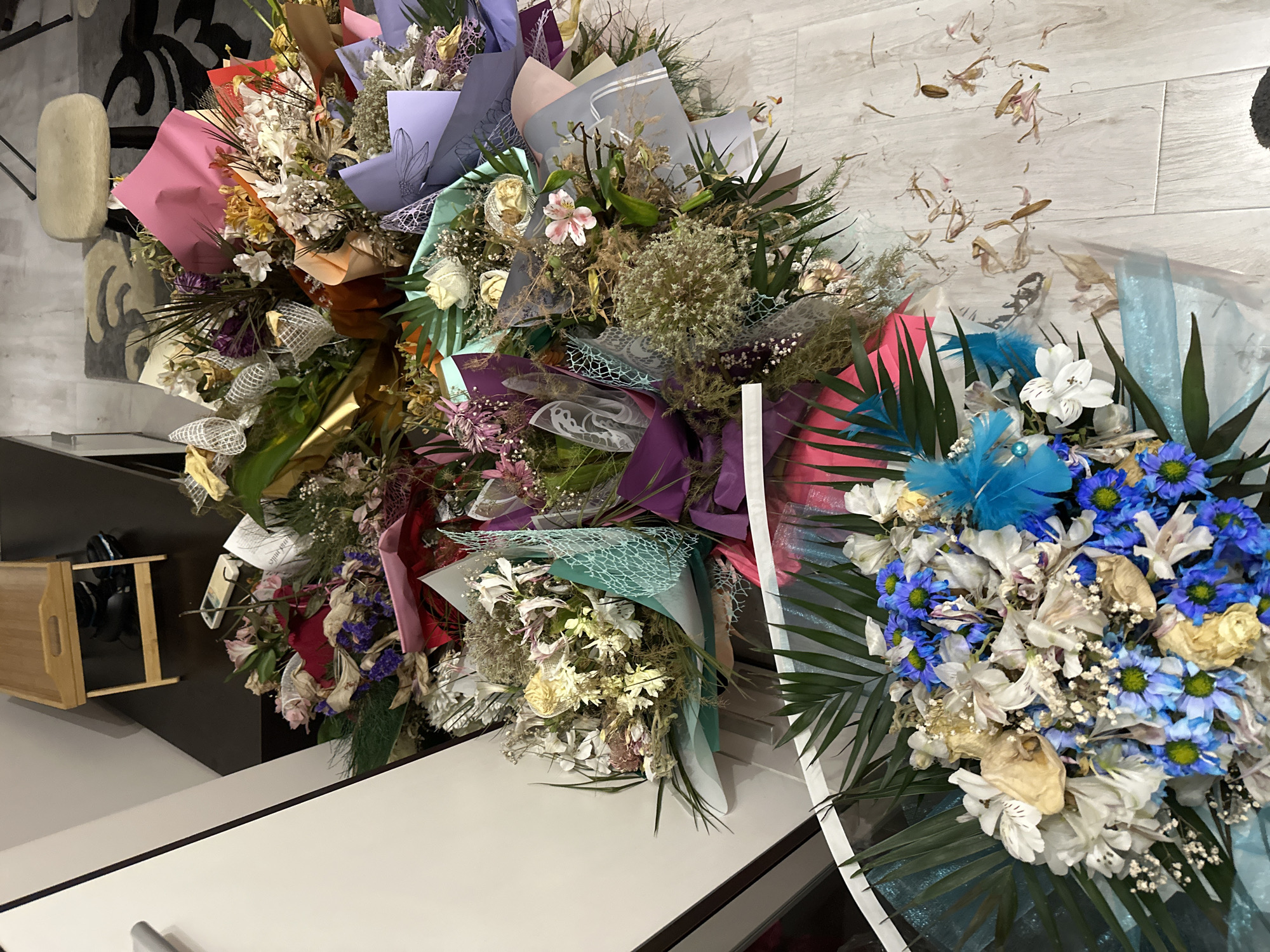 A top-down interior shot captured in Unknown, Unknown, displays a collection of approximately 15 or more wilted and dried floral bouquets arranged on a light-colored wooden or laminate floor. The bouquets, varying in size and composition, feature faded flowers in shades of white, cream, pink, purple, blue, yellow, and red, intermingled with dried green foliage. Each bouquet is individually wrapped in various materials, including matte and metallic papers in colors such as hot pink, orange, light blue, mint green, purple, light grey, gold, and red. Several wrappings incorporate decorative mesh fabrics in white, teal, and grey patterns. One bouquet in the foreground exhibits prominent blue chrysanthemums, white alstroemeria, and green palm fronds, accented with a blue feather. Scattered dried petals and floral debris are visible on the floor, particularly in the lower right portion of the image.
The background reveals a white wall or cabinet with a silver handle on the left. In the upper left, a light-colored wooden serving tray rests on a dark surface, with dark objects resembling shoes or electronics partially visible beneath it. Towards the upper right, a small, light-colored stool with a shaggy top is situated on a dark floor with a patterned grey and black rug. Further into the background, a remote control and various papers are visible on a dark surface, along with a yellow cloth or garment draped over an unseen item. No individuals are present in the scene.