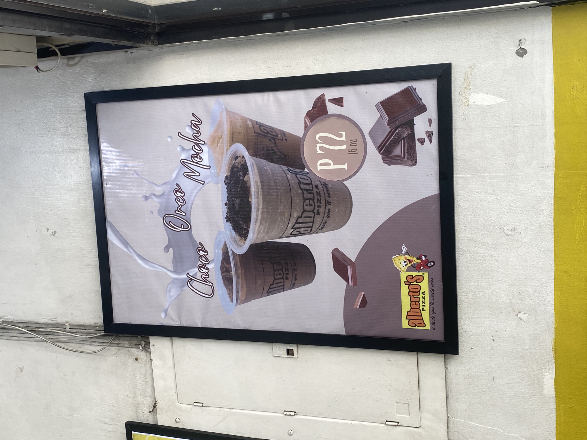 A framed advertisement poster is prominently displayed on an off-white, textured wall, likely in Davao City, Philippines. The poster features three clear plastic cups filled with iced beverages, set against a light background with a white splash resembling milk or cream. The central cup contains a dark brown drink topped with crushed cookies, while the others appear to be a lighter brown or caramel color.
Text on the left side of the poster reads "Choco Oreo Mocha" in a stylized, cursive font. A circular emblem on the right displays the price "P 72" and size "16 oz," indicating Philippine Pesos. Chocolate bar pieces are scattered around this emblem and below the drinks. The brand "Alberto's PIZZA" is visible on the cups and on a yellow logo at the bottom right of the poster, accompanied by the tagline "a taste you'll surely miss..." The logo itself depicts a cartoon character, possibly a chef or delivery person, holding an item.
The wall shows signs of wear and tear, with some visible cracks and chipped paint. To the right of the poster, a vertical section of the wall is painted bright yellow. Above the poster, a dark metal frame structure is visible, along with some exposed wires. Below the poster, there is a white panel with hinges, suggesting a counter or closed storage area. The overall scene appears to be an outdoor or semi-sheltered area, possibly a food stall or a part of a building exterior.