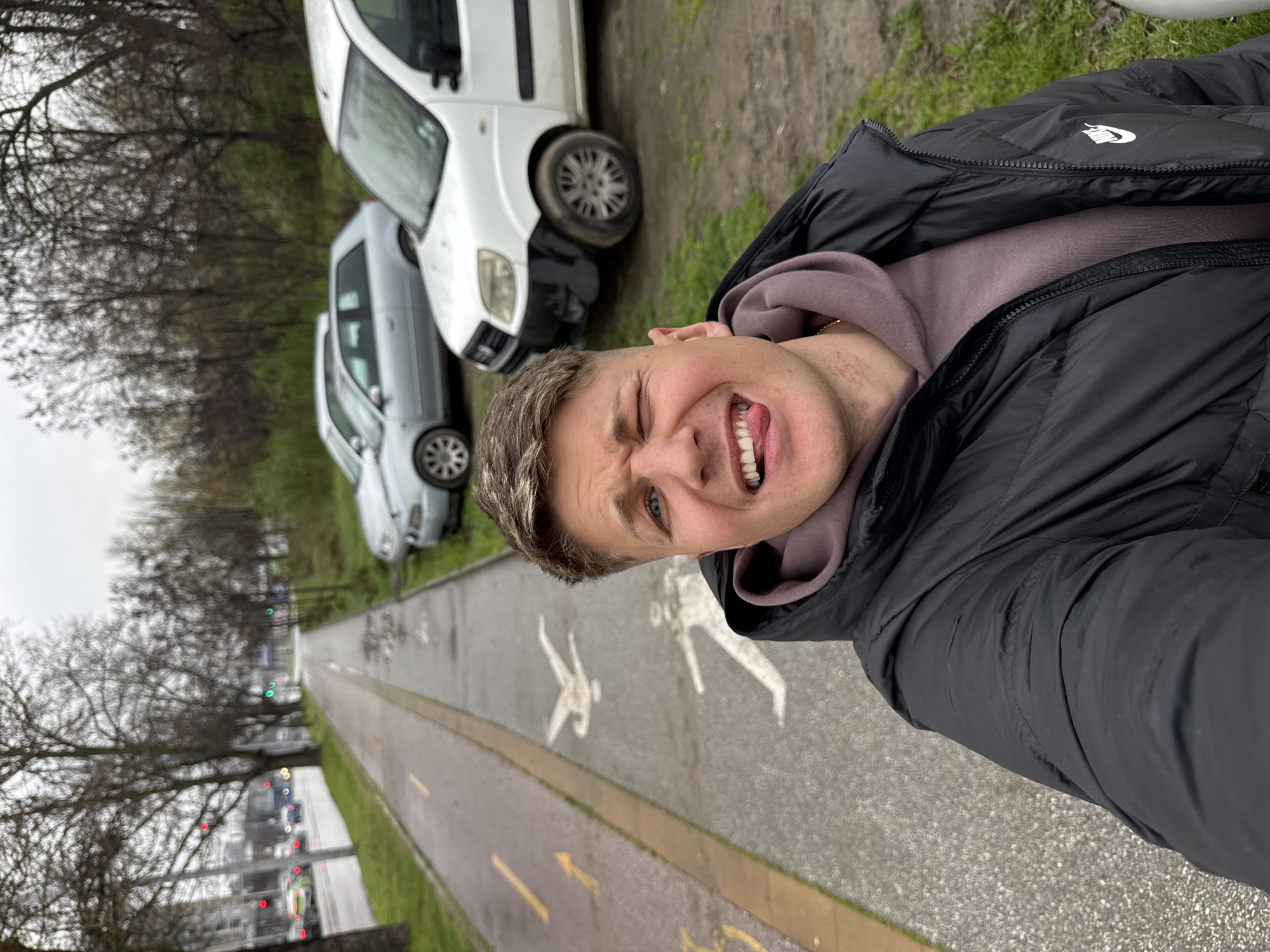 A person is taking a selfie in Plovdiv, Bulgaria, along a combined pedestrian and bicycle path marked with yellow lines and symbols indicating pedestrian and bicycle lanes. They are wearing a black puffer jacket over a mauve hoodie. Two parked white cars are visible to the right of the path. On the left side, bare trees line the path, and traffic lights are visible in the distance.