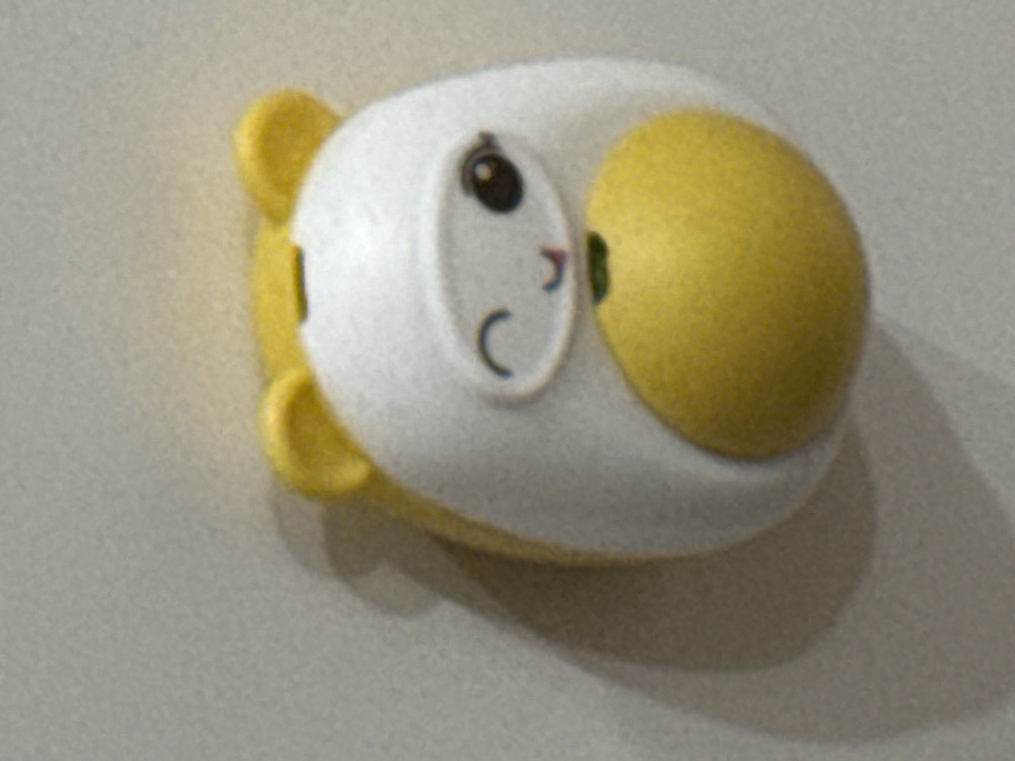 The image shows a close-up, slightly blurred view of a yellow and white object, resembling a toy or a decorative item, mounted on a light-colored, textured surface. The object has two small, rounded yellow protrusions at the top, which appear to be ears. The main body of the object is predominantly white, with a large, circular yellow section at the bottom. A cartoonish face is visible on the white part, featuring one dark, circular eye and a winking eye with an eyelash. A small, curved mouth with a pinkish hue is also depicted. There is a hint of green just below the mouth. The object is casting a soft shadow on the surface it is mounted on. The background is a uniform, neutral tone, showing signs of texture and possibly being a wall or a similar surface. There is no indication of Saratov, Russia, or any urban environment within the provided image.