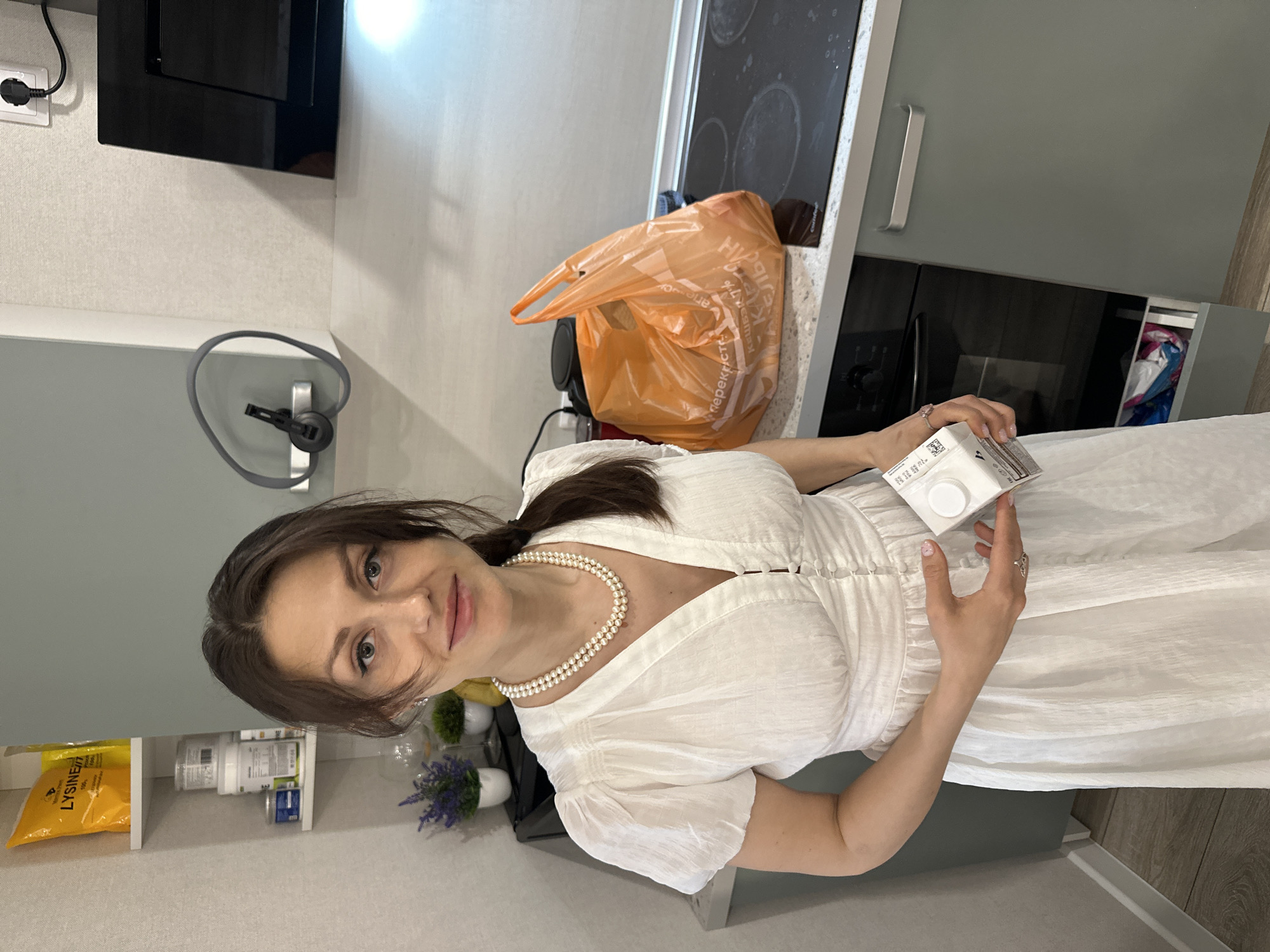 A woman in a white dress and pearl necklace stands in a kitchen in Murino, Russia, holding a white carton. An orange plastic shopping bag sits on the counter behind her, near a black stovetop. A shelf with supplements and other items is visible to her left. A black microwave is mounted on the wall.