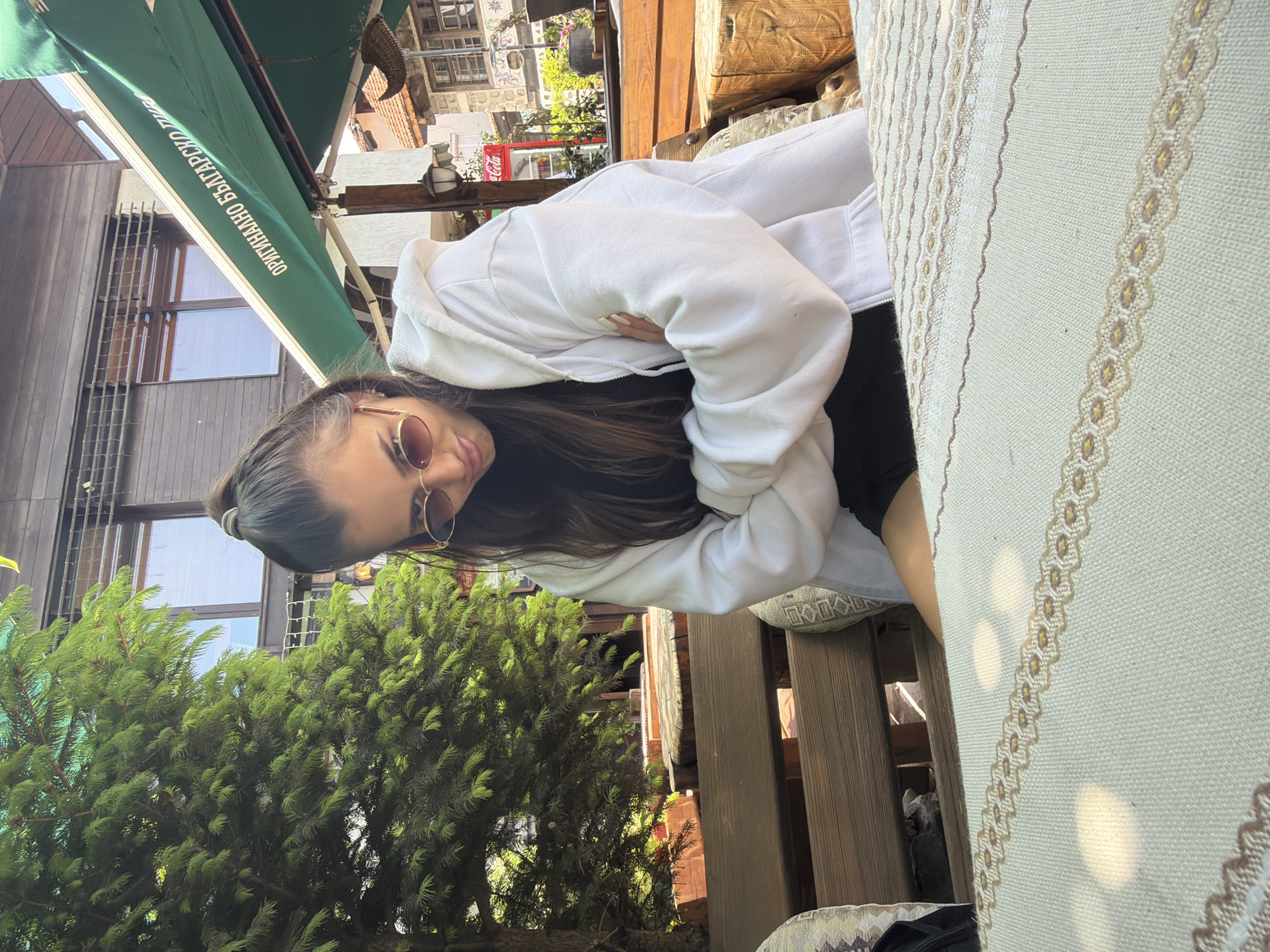 A young adult female is seated outdoors at a table, viewed from a low-angle perspective. She has long, dark hair pulled back into a ponytail and is wearing oval, reddish-brown tinted sunglasses with thin gold-colored frames. She is dressed in a white, zippered hoodie over a dark top, with her arms crossed over her chest. Her gaze is directed downward and slightly to her left. Her left leg, visible from mid-thigh, suggests she is wearing dark shorts or a short skirt.
In the immediate foreground, a light-colored woven tablecloth with intricate beige and brown geometric patterns covers the table. Irregular patches of sunlight create bright reflections on its surface.
Behind the woman, a wooden bench and parts of other wooden furniture are visible. To the far left, a dense green coniferous tree stands. In the mid-ground, a two-story building with dark horizontal wooden siding and multiple windows reflecting the sky is present. Above a section of this building, a green awning extends, displaying white Cyrillic text that reads "ОРИГИНАЛНО БЪЛГАРСКО ПИВО" (ORIGINAL BULGARIAN BEER). Below the awning and to the right of the tree, a red Coca-Cola cooler/refrigerator is visible next to an old-fashioned black street lamp attached to a wooden post. Further to the right in the background, segments of traditional buildings with orange-red tiled roofs and stone walls can be seen, along with a decorative wicker object hanging from a structure. The scene is brightly illuminated by natural sunlight, indicating daytime.