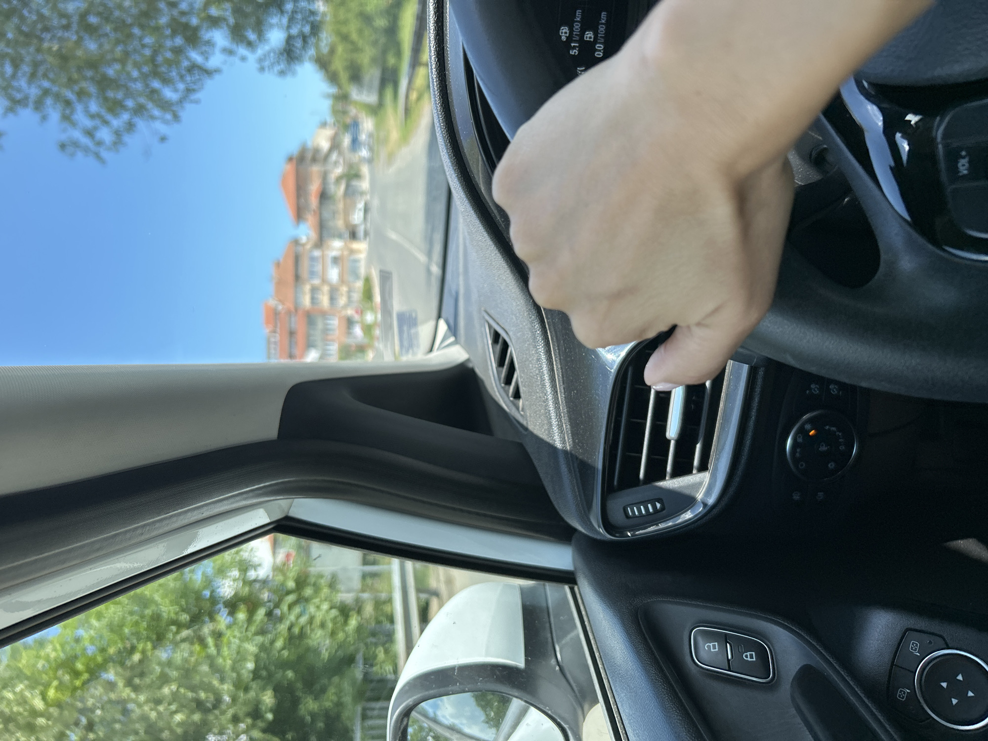 The image shows a first-person perspective from inside a car driving on a sunny day in Aheloy, Bulgaria. The driver's right hand is visible on the steering wheel. The car's dashboard displays a digital readout showing "5.1 1/100 km" and "0.0 1/100 km". Outside the car, a road is visible, lined with trees and buildings with orange roofs. The sky is clear and blue. The overall scene depicts a typical drive through a town on a bright summer day.