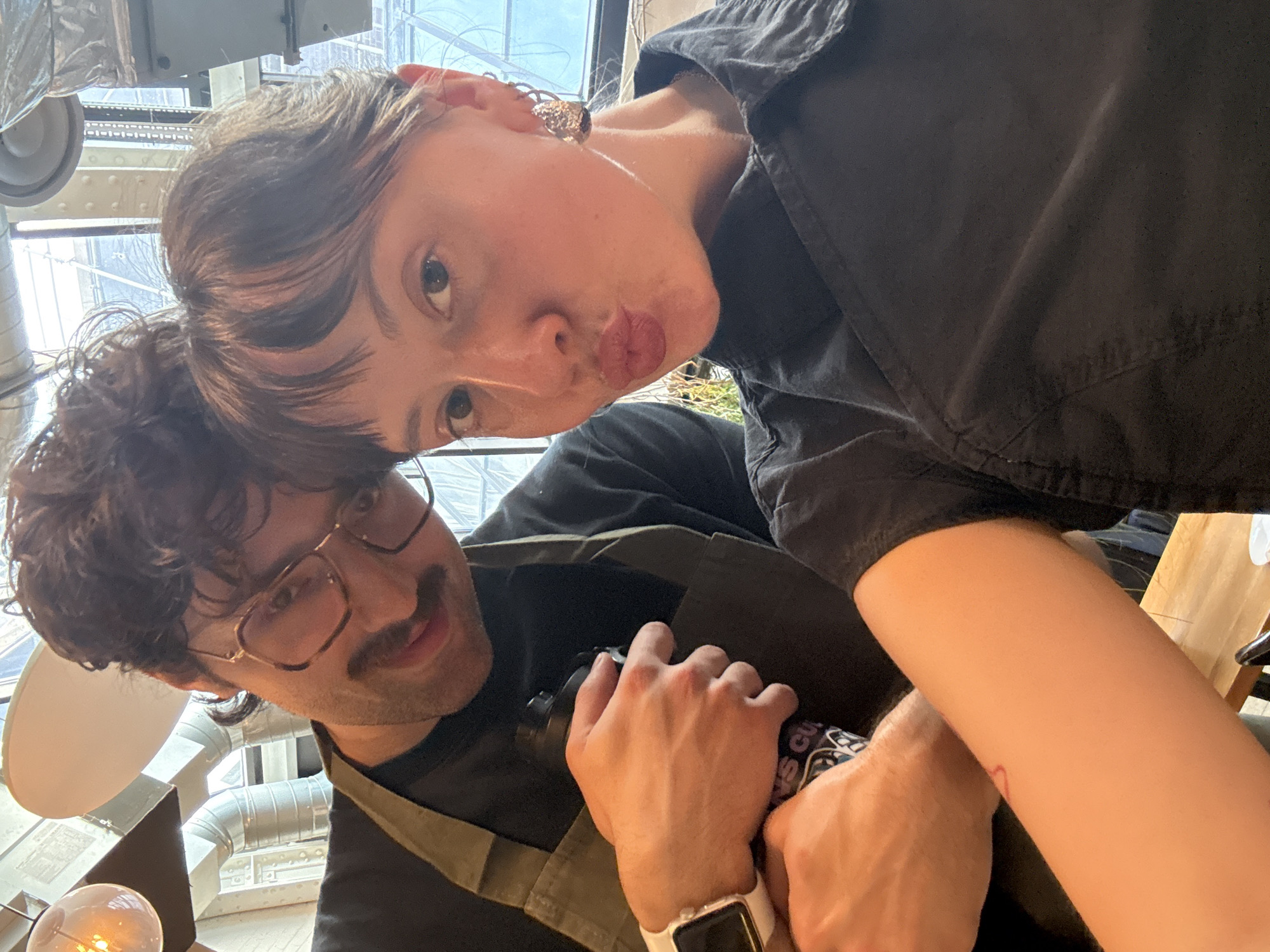 This is a close-up, upward-angled selfie featuring two individuals in an indoor setting, likely in London, United Kingdom. The image is significantly tilted to the right.
In the foreground, an individual with short, dark, textured hair and fair skin is seen. They are wearing a dark, possibly black, button-up shirt and an intricate, coiled metal earring on their right ear. Their lips are pursed in a playful "kissing" expression as they look directly at the camera. A faint red line, possibly a small tattoo, is visible on their inner left forearm.
Below and slightly behind the first individual is a male with dark, curly hair, a mustache, and a slight beard shadow. He wears rectangular glasses and a dark t-shirt under what appears to be a light brown or olive green apron or strap. He looks towards the camera with a subtle smile. His left wrist bears a white Apple Watch. He is holding a dark bottle or thermos with both hands; text visible on the bottle includes "NEWS CULT" alongside graphic designs.
The background suggests a modern, industrial-style interior, characterized by exposed metal ductwork and structural elements, and large windows that let in ample natural light, indicating it is daytime. A light fixture with an exposed spherical bulb is visible in the lower left corner.
The overall mood appears lighthearted and intimate, possibly capturing a shared moment between friends or a couple.