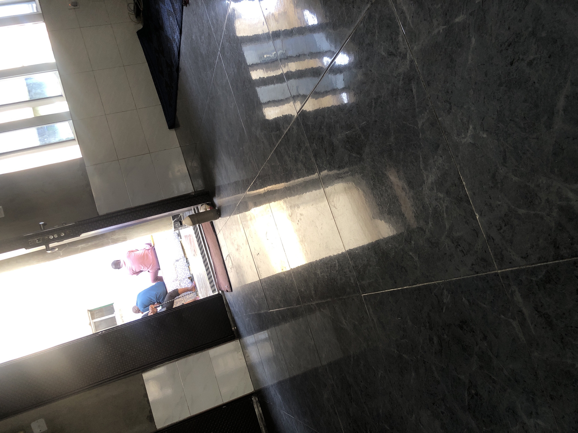 The image shows a brightly lit interior with dark, reflective floor tiles. In the background, visible through an open doorway, three individuals are seated. One person, wearing a blue garment, is facing away from the camera and appears to be holding a cane. To their right, another individual, dressed in pink, is also facing away. A third person, wearing a white and pink outfit, is seated between them. The doorway leads to an outdoor area with a white tiled bench. The overall impression is of a communal space, possibly a religious or social gathering area, located in Ado, Nigeria. Sunlight streams into the room from the doorway and through a window with multiple panes, creating reflections on the polished floor. A dark rug is partially visible in the upper right portion of the image, near the tiled wall. A brown plastic bottle is standing near the doorway on the right side.