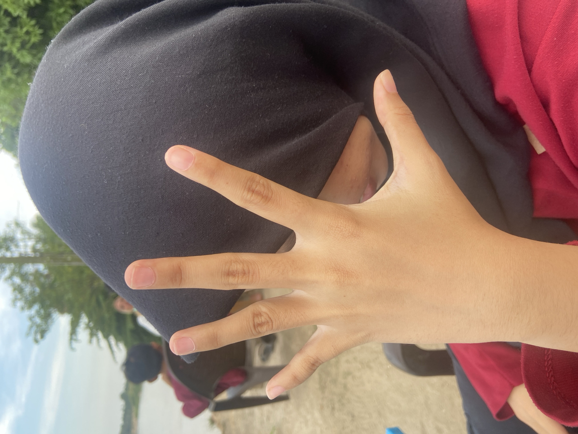 The media file shows a close-up of a person's hand with their fingers spread out, partially covering their face, which is obscured by a black head covering. The person is wearing a red long-sleeved shirt. In the background, out of focus, trees and a body of water are visible, suggesting an outdoor setting. There are other people in the background, one of whom appears to be wearing a black head covering and a maroon shirt. The scene seems to be taking place during the daytime, with natural lighting. The image is taken from a slightly downward angle, looking towards the person's hand. The focus is on the hand and the black head covering. No text is visible in the media file.