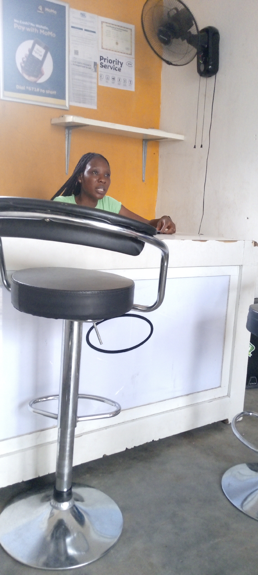 The image depicts the interior of what appears to be a service-oriented establishment in Zango-ndaji, Nigeria. A woman with braided hair, wearing a green t-shirt, stands behind a counter with a white facade. She appears to be in conversation. The counter is positioned in front of a yellow wall. Above the counter are several framed signs and advertisements, including one that says "Pay with MoMo". A shelf is positioned beneath the signs. To the right, a black wall-mounted fan is visible. In the foreground are two chrome bar stools with black seats. The floor is gray concrete.