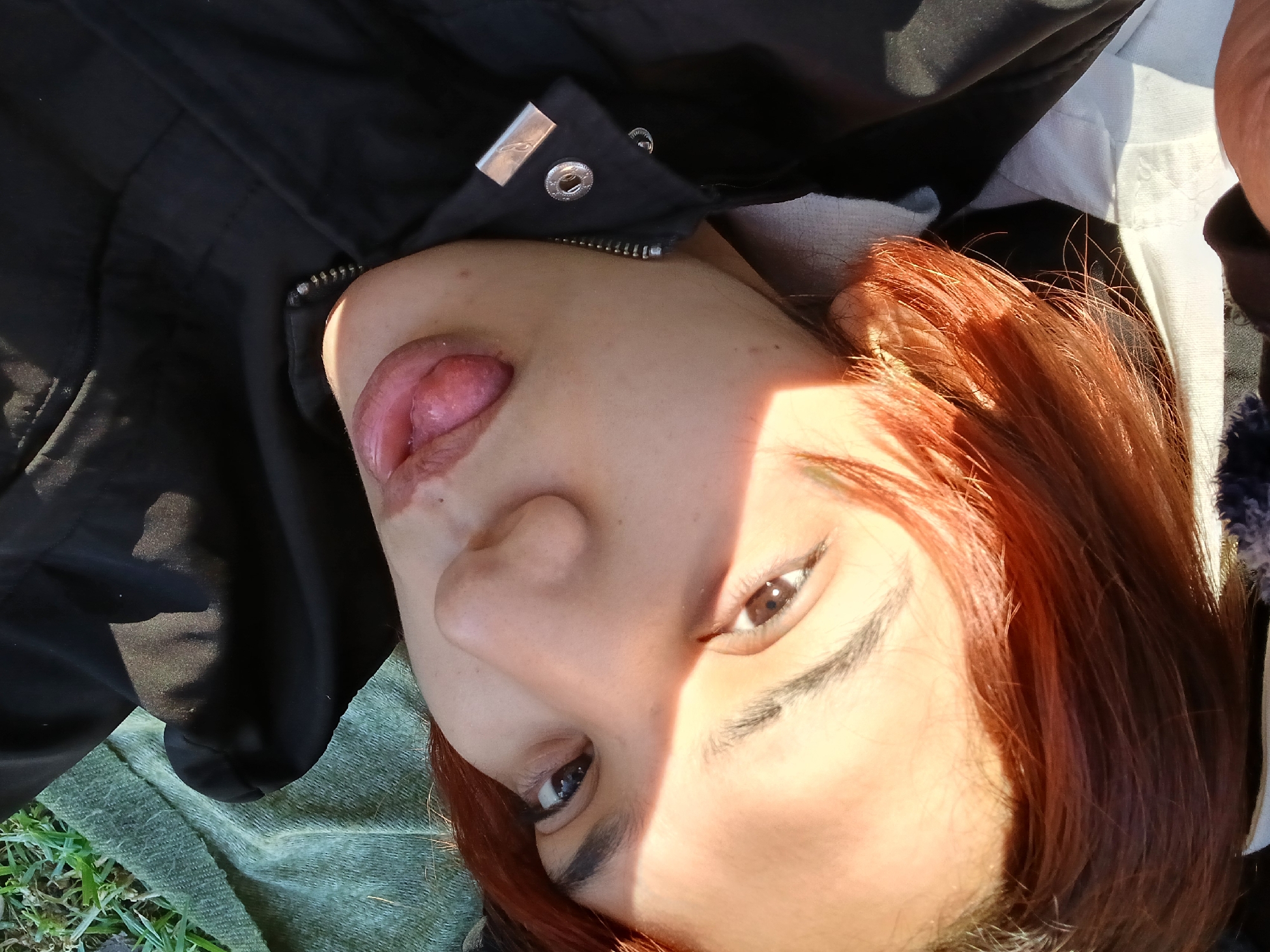 A person is lying on their back in an outdoor setting. The person has red hair, fair skin, and is wearing a black jacket with metal snaps near the collar. The person has their tongue sticking out. The person is lying on green fabric or grass. There is sunlight illuminating one side of the person's face. The location is possibly a park or grassy area, but the specific location in Buenos Aires, Argentina is indeterminable.