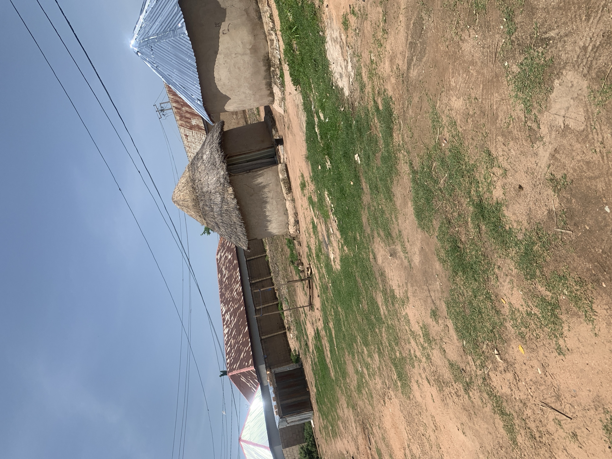 This image depicts a scene in Jauro Yinu, Nigeria, showcasing a collection of traditional dwellings. The foreground consists of a dirt path with sparse patches of green grass. In the mid-ground, several circular structures with mud walls are visible. One structure has a thatched roof, while another is topped with a shiny, blue, corrugated metal roof. Behind these, a longer building with a rusty, corrugated metal roof is present, bordered by a low wall. Another dwelling with a light-colored, corrugated metal roof is situated to the far left. Overhead, power lines stretch across a clear blue sky with scattered clouds. The overall impression is that of a rural or semi-rural residential area.